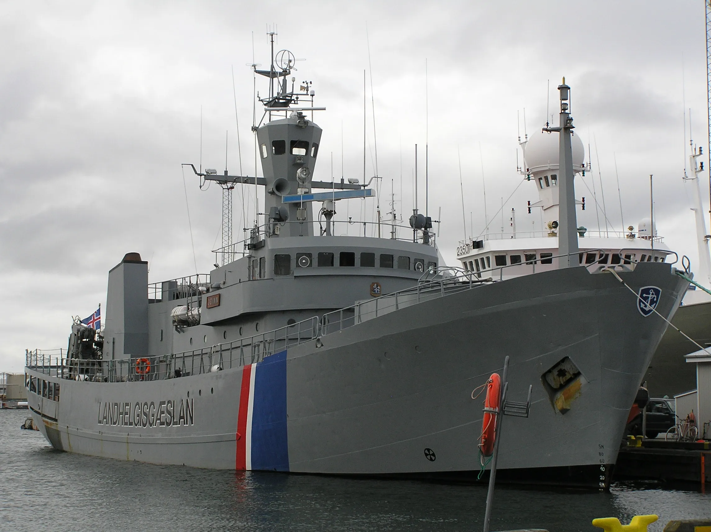 Patrullera gris de la Guardia Costera de Islandia Óðinn atracado en un puerto, mostrando el casco con el texto LANDHELGISGÆSLAN y una franja roja y azul, la superestructura, el mástil y las antenas bajo un cielo nublado