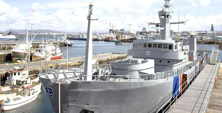 Gran embarcación gris de la Guardia Costera atracada en un muelle con barcos más pequeños y un paisaje urbano de fondo