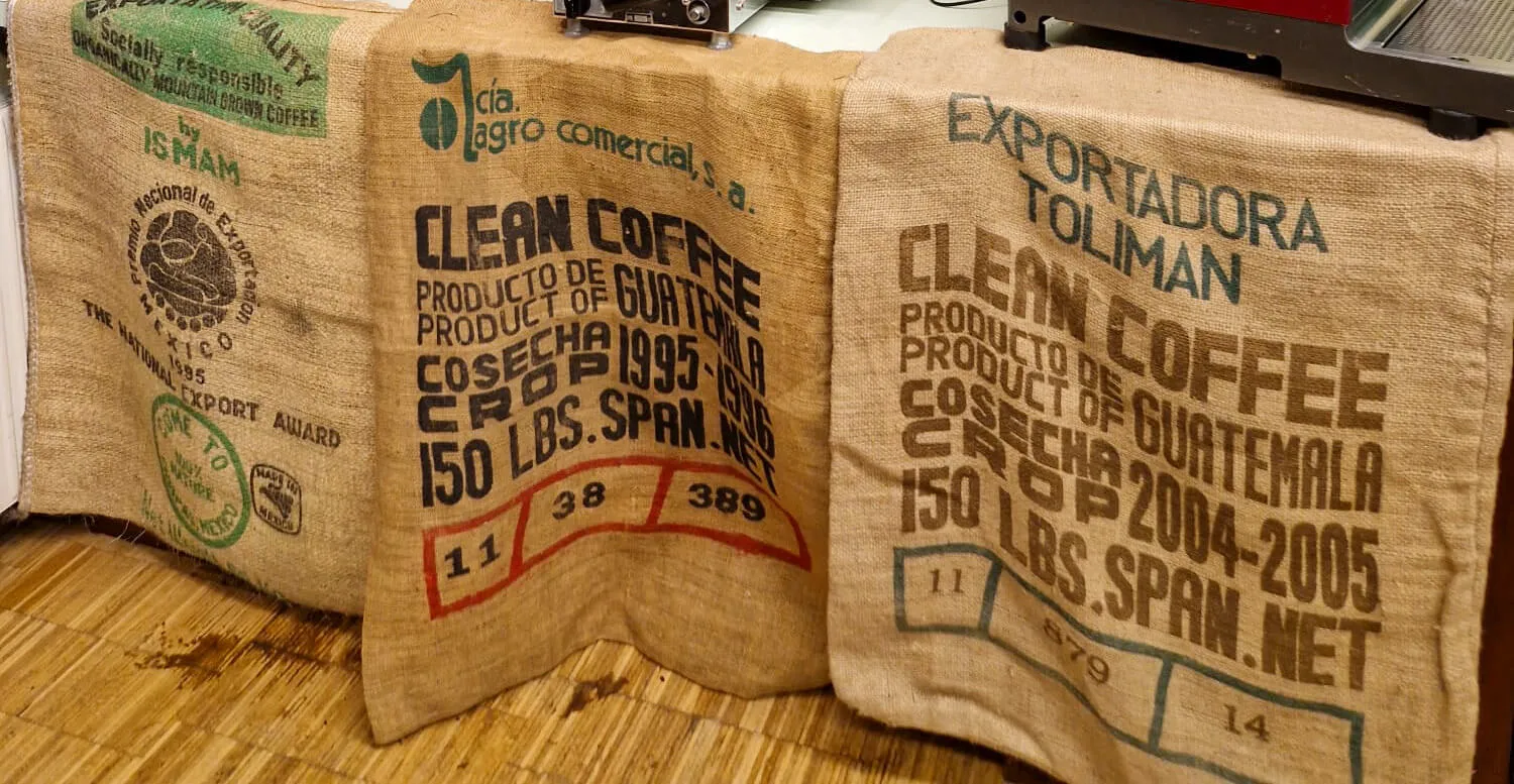 drie koffiezakken van jute met tekst en logo's op een houten ondergrond