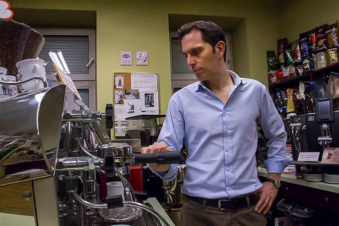Een man die een espressomachine bedient in een koffiemuseum met planken vol koffieproducten en -apparatuur op de achtergrond