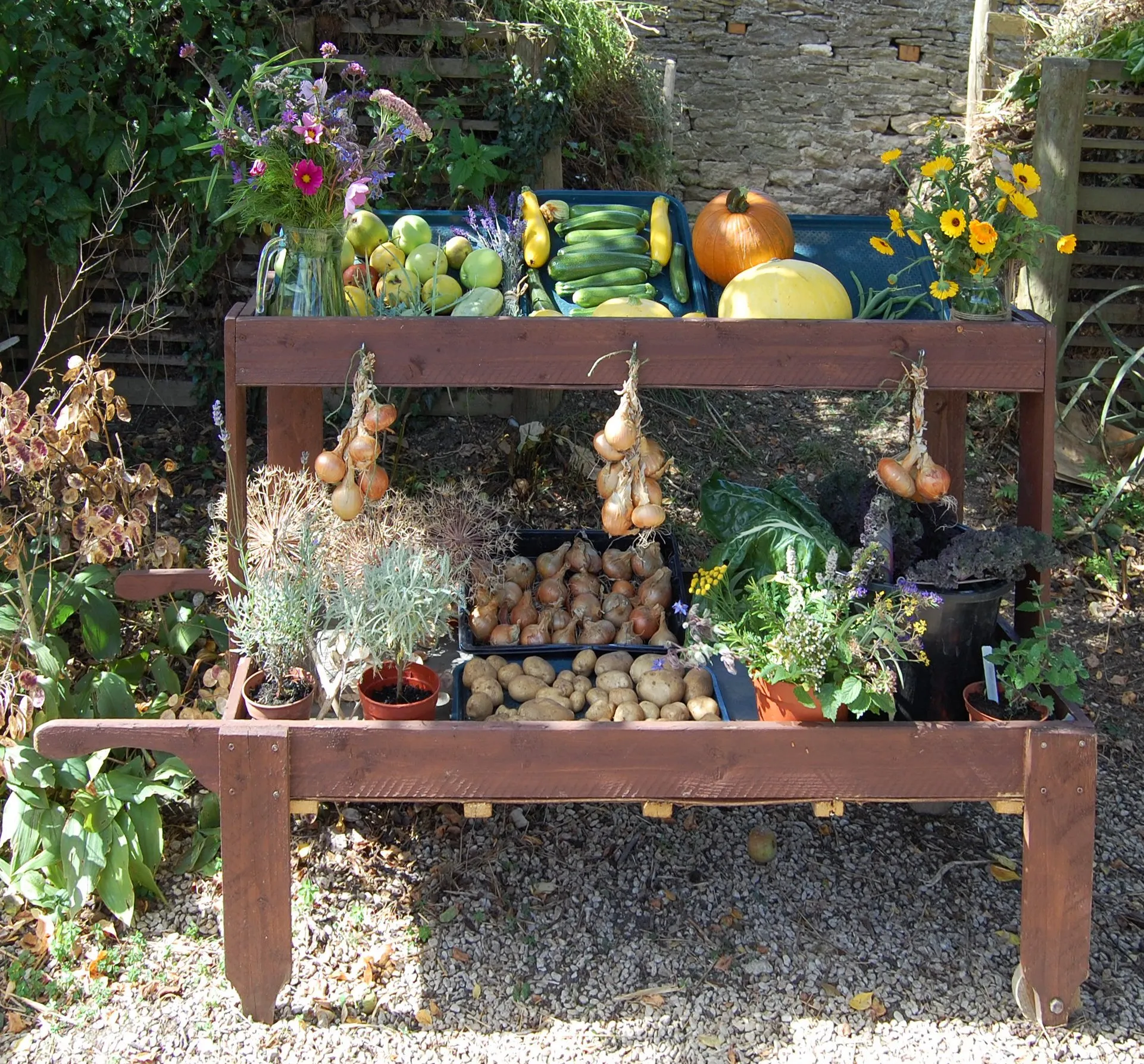 Houten verpotbank buiten met daarop geoogste producten zoals appels, courgettes, pompoenen, hangende uien, kratten met aardappelen en potkruiden, met bloemen en een stenen muur op de achtergrond.