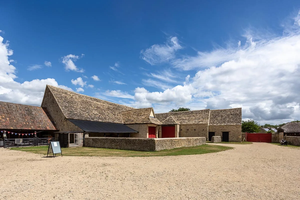Stenen boerderijgebouwen met pannendaken en rode deuren rondom een grindplaats onder een blauwe lucht met wolken bij Cogges Manor Farm