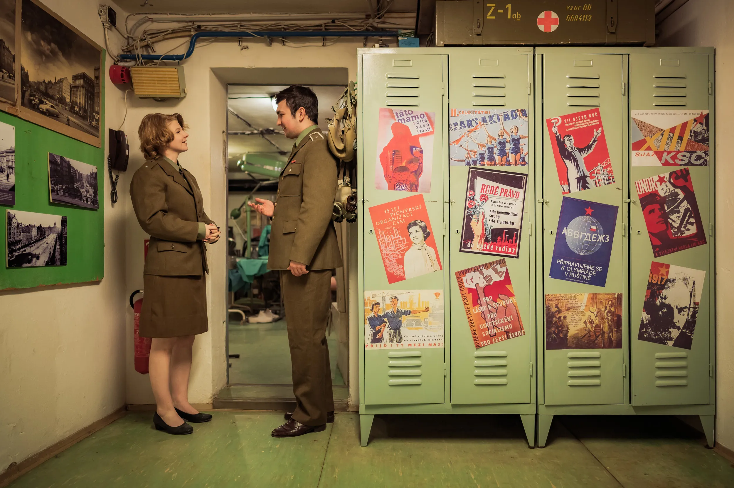 Two people in Cold War-era clothing standing in a hallway with green lockers covered in propaganda posters and a display board with historical photographs