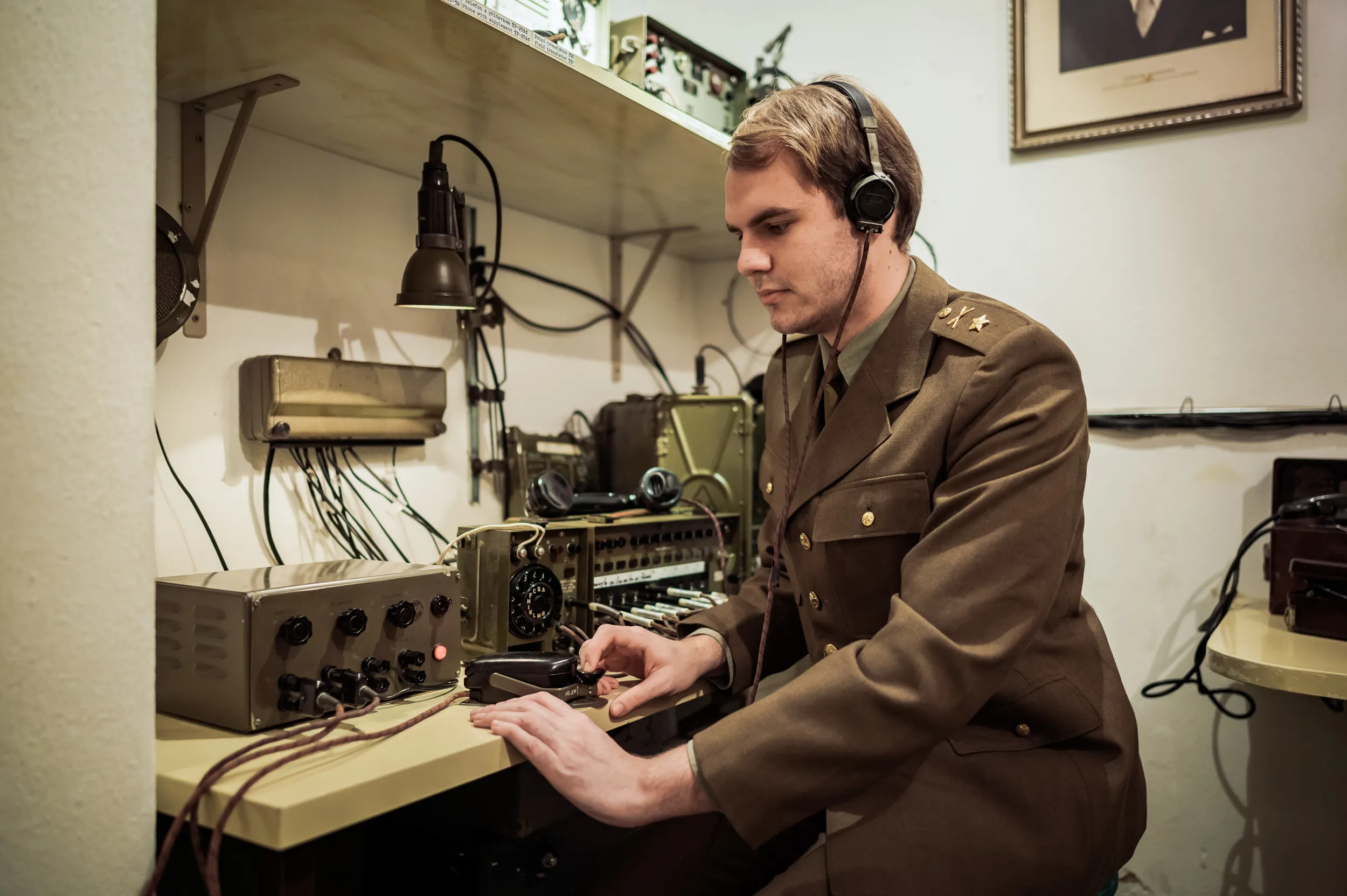 Man in military uniform operating vintage radio equipment in a dimly lit room with wires and old electronics