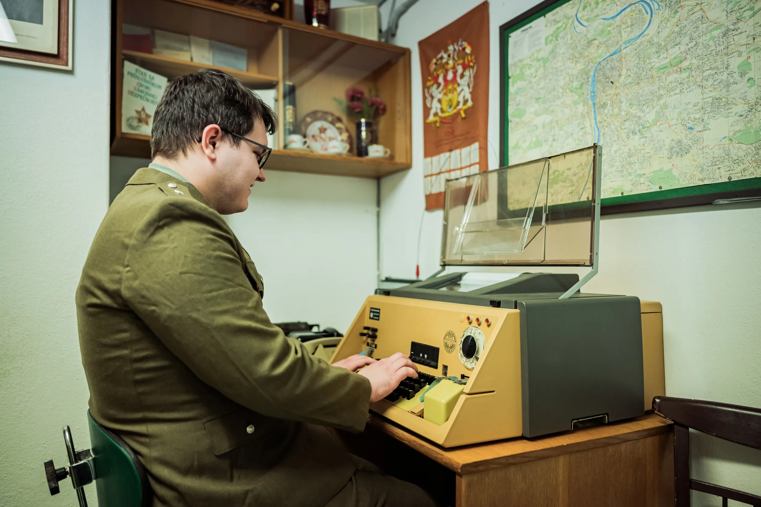 Man in a green military uniform operating a yellow and gray vintage communication machine in a room with a map on the wall