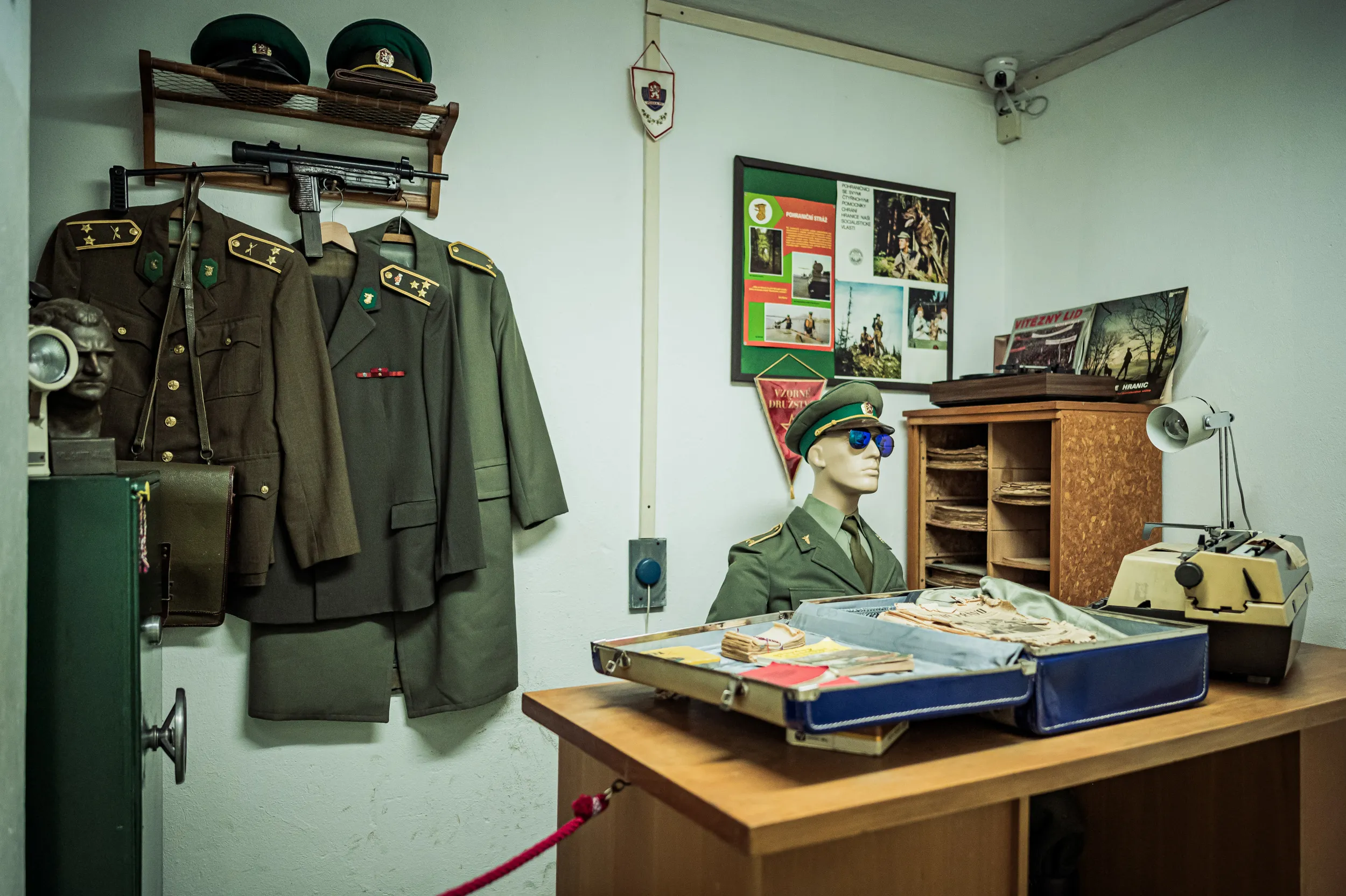 Military uniforms hanging on a wall, a mannequin in a green uniform, and a display case with documents and equipment on a wooden table