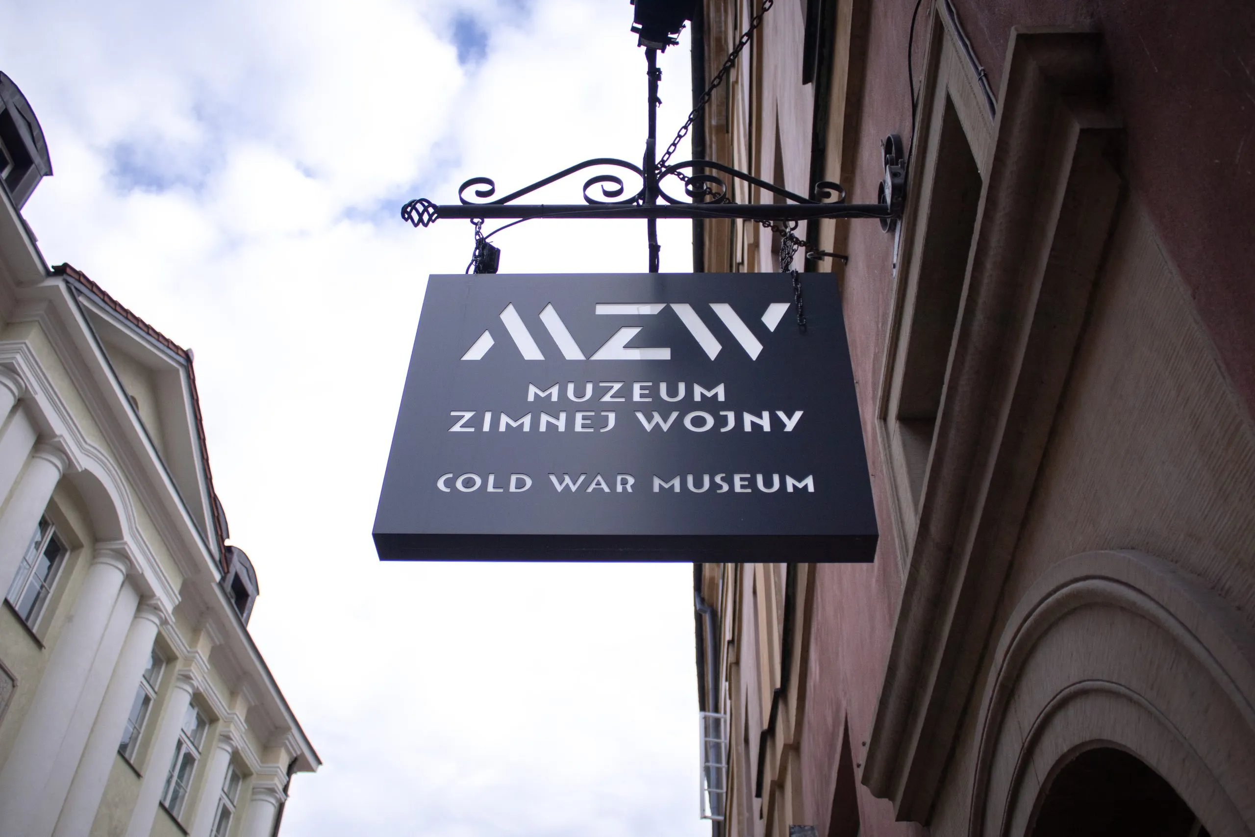 Cartel negro con texto blanco 'MZ MUSEUM ZIMNEJ WOJNY COLD WAR MUSEUM' colgado de un soporte decorativo en la fachada de un edificio.