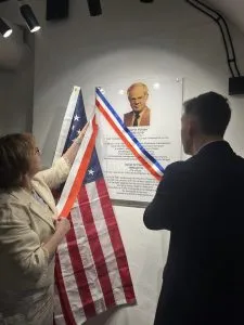 Dos personas de pie frente a una exhibición en la pared con una bandera estadounidense y un retrato de un hombre en traje.