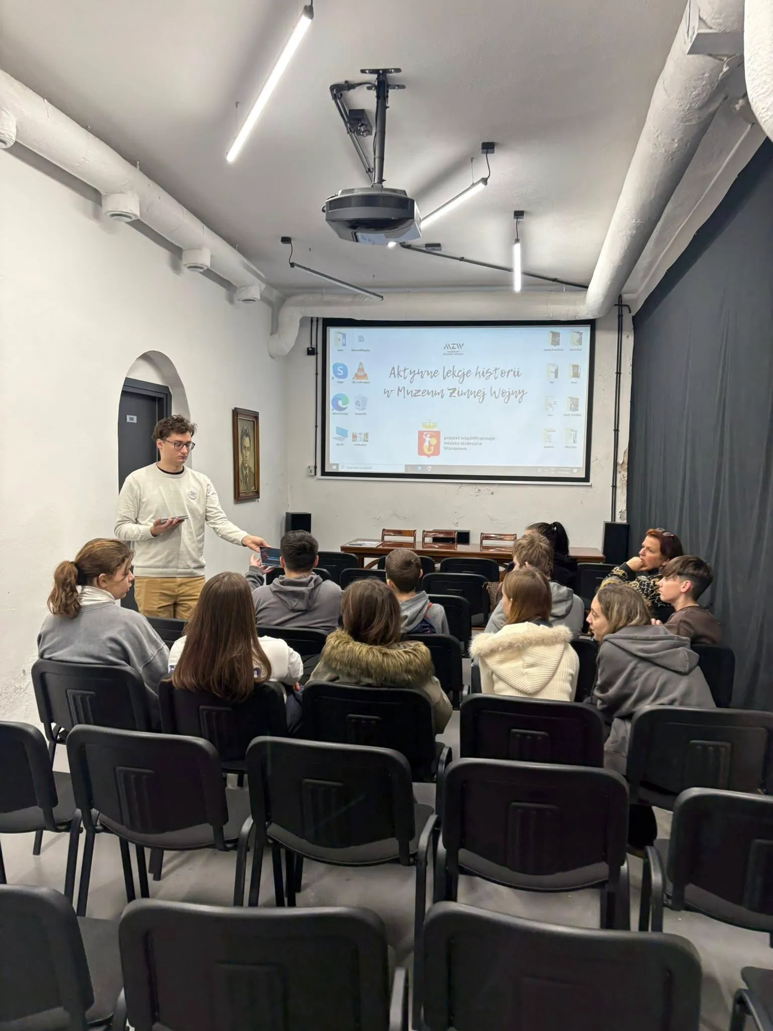 Een spreker die tot een groep studenten spreekt in een collegezaal met een projectiescherm waarop een dia met Poolse tekst en logo's wordt getoond