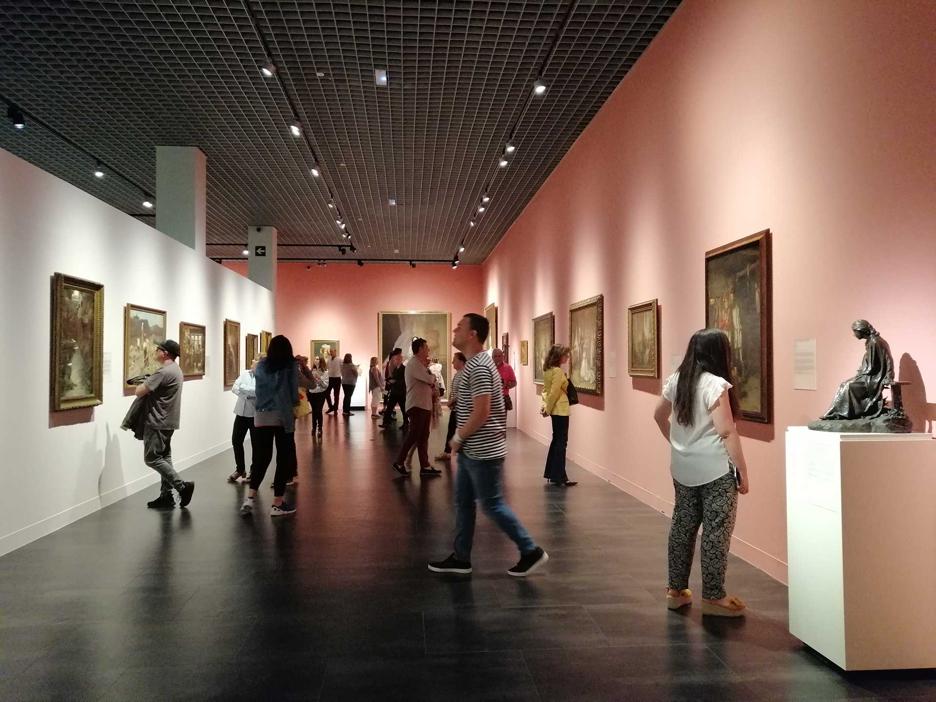 Museum gallery interior with visitors viewing framed paintings on pink walls and a bronze sculpture on a white pedestal under a dark grid ceiling