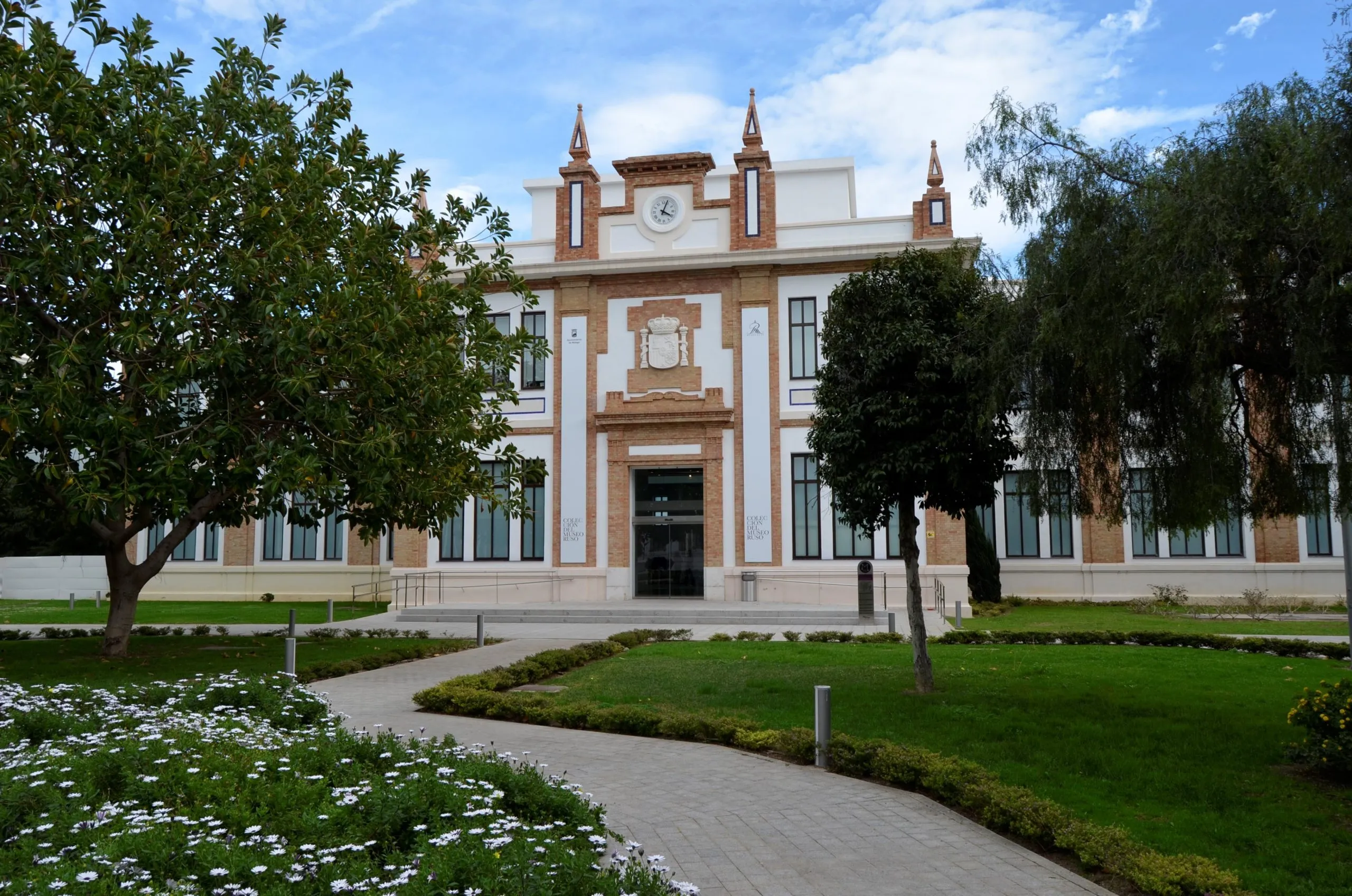 White and terracotta facade of the Colección del Museo Ruso Málaga building with decorative towers, central entrance, and a winding garden path flanked by trees and flower beds in the foreground.