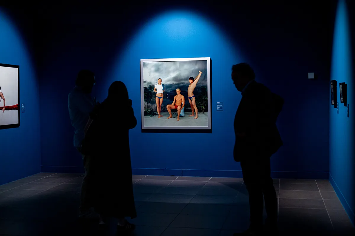 Silhouetted visitors stand in a dark gallery with deep blue walls, viewing spotlit framed photographic artworks including a central image of three men in a landscape.