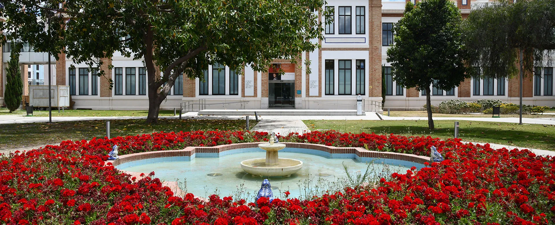 Wide view of a multi-story building with a circular fountain surrounded by a dense bed of red flowers in the foreground courtyard.