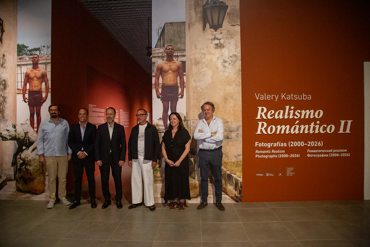 Six people posing in front of a large exhibition wall displaying Valery Katsuba Realismo Romantico II photography exhibition at Coleccion del Museo Ruso Malaga