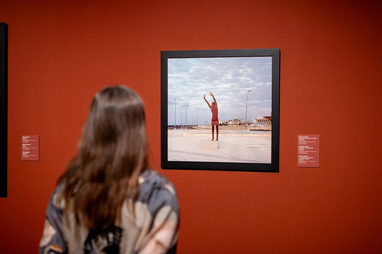 A visitor with long brown hair viewed from behind looks at a framed photograph of a figure in red on a deep red gallery wall at the Colección del Museo Ruso Málaga