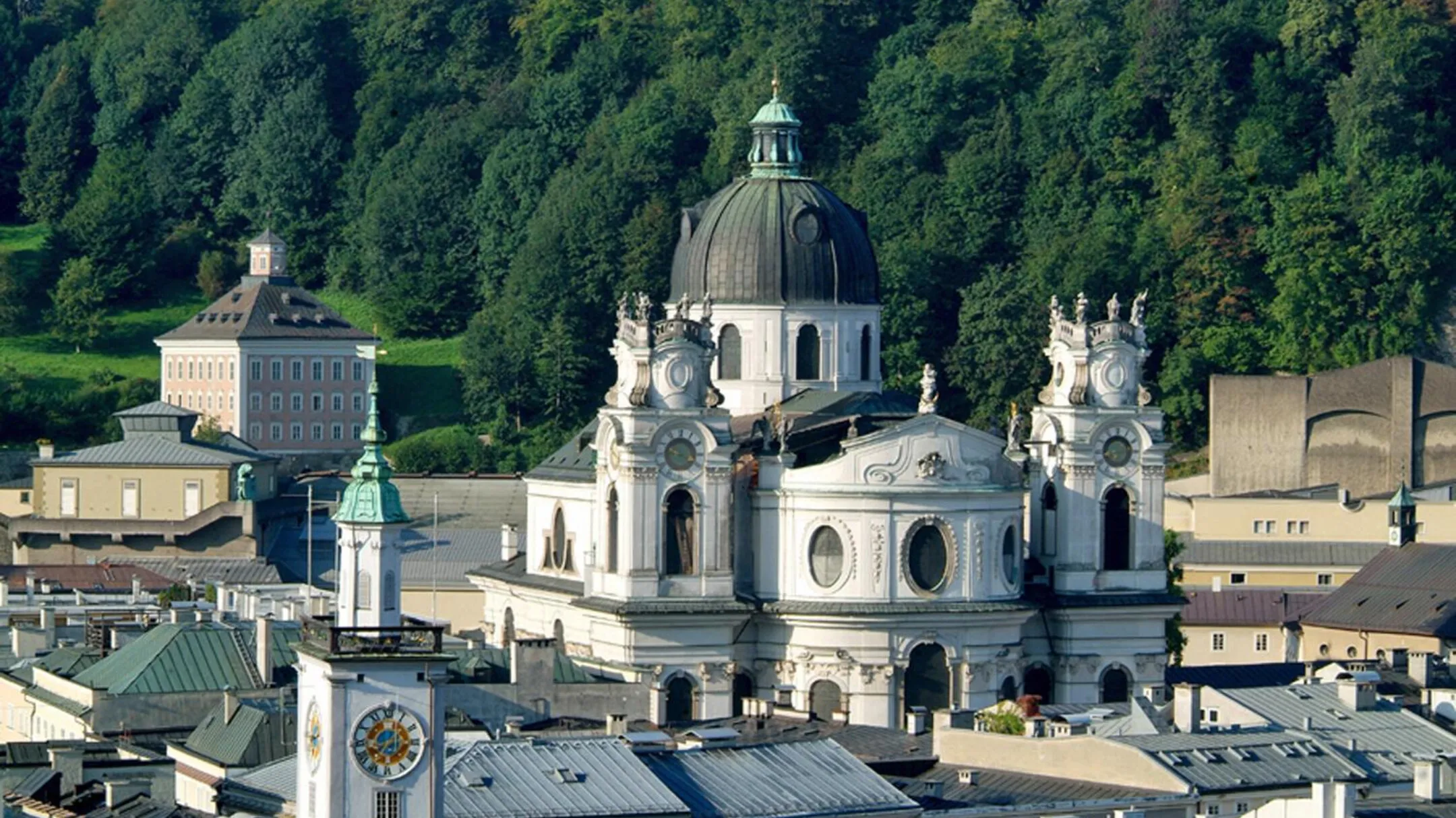 Vista aérea de la blanca iglesia barroca Kollegienkirche con su gran cúpula y torres gemelas, rodeada por los tejados de la ciudad de Salzburgo y una colina boscosa