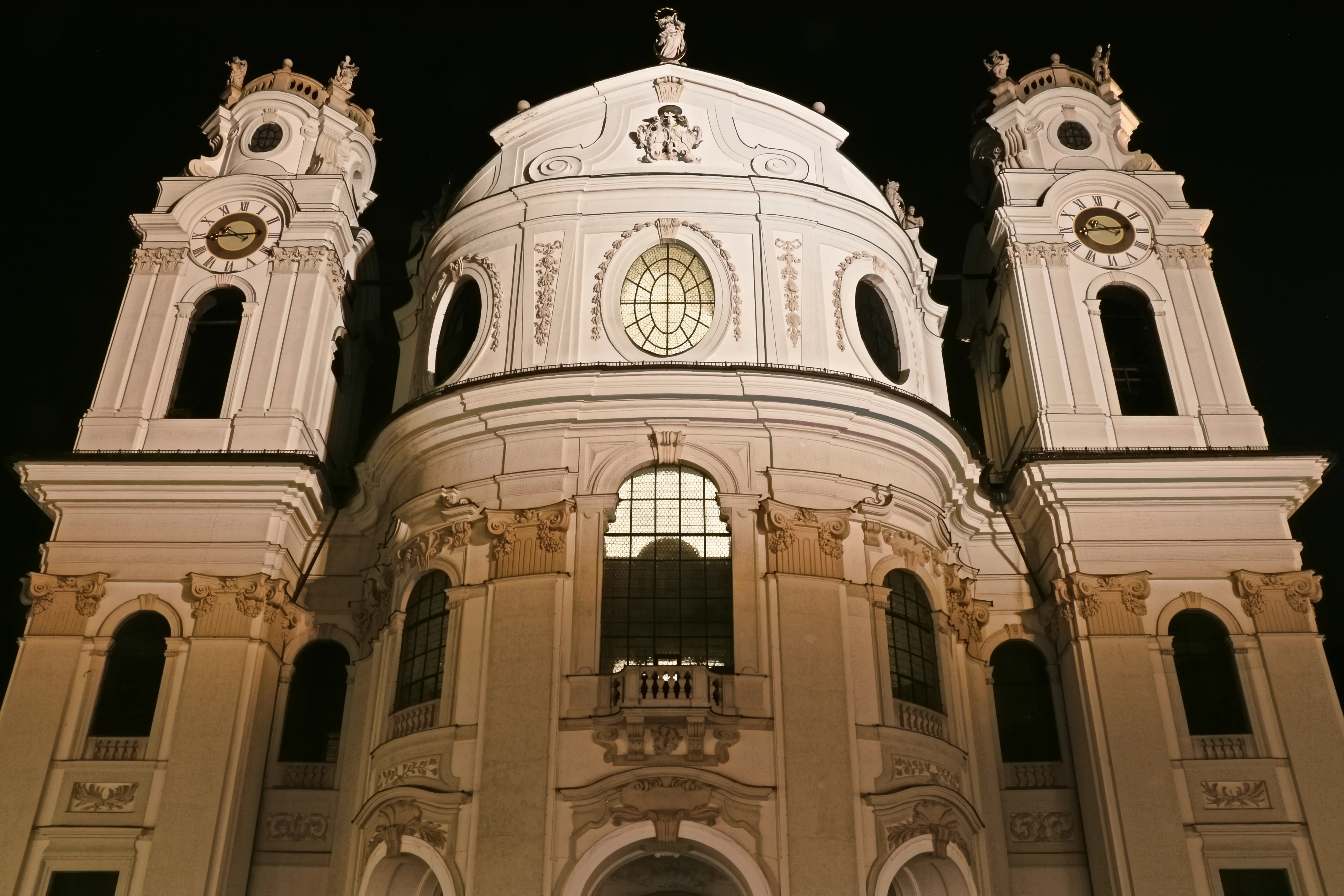 Fachada de iglesia barroca blanca de noche con dos torres de campana iluminadas flanqueando una cúpula central, ventanas arqueadas y detalles arquitectónicos ornamentados sobre un cielo oscuro