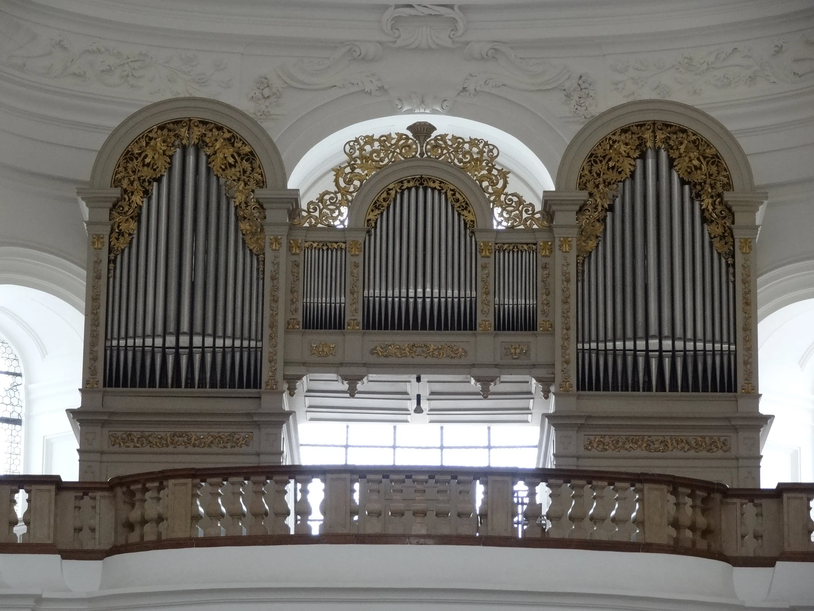 Gran órgano de tubos con ornamentos dorados colocado en tres nichos arqueados blancos sobre una barandilla de madera en el interior de una iglesia barroca.