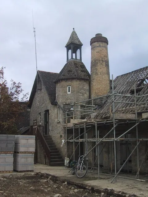 Edificio de piedra de Combe Mill con una torre coronada por una cúpula, una alta chimenea de ladrillo y andamios cubriendo parte del techo durante las obras de restauración, con una bicicleta aparcada cerca.