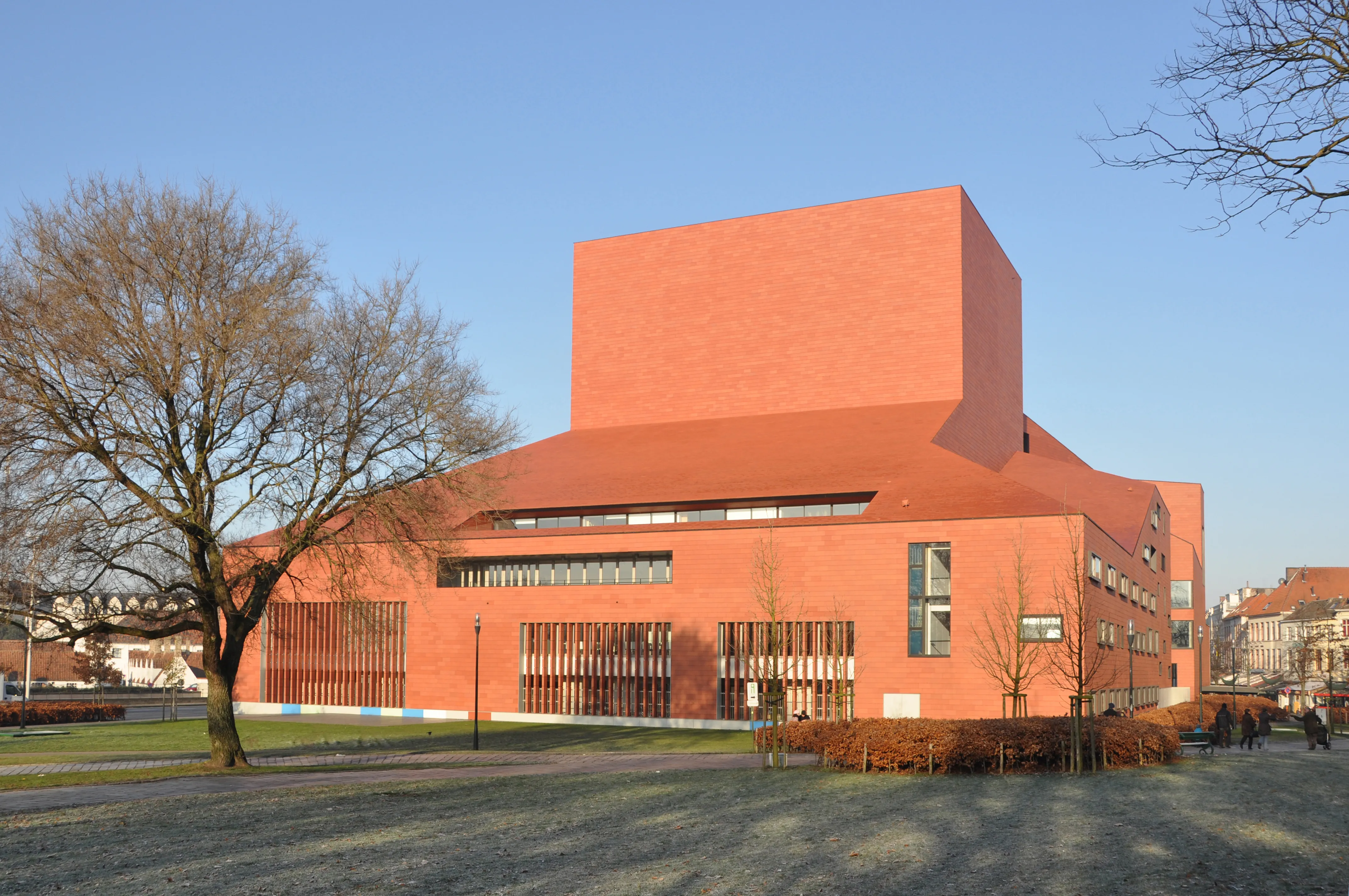 Vista exterior del Concertgebouw Brugge, una gran sala de conciertos de ladrillo rojo terracota con una distintiva estructura superior cúbica, sobre un cielo azul despejado con árboles desnudos en primer plano.