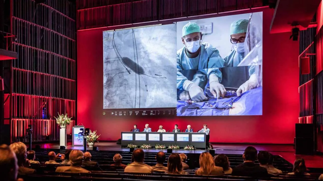 Panel de ponentes en el escenario de un gran auditorio con paredes rojas, con una pantalla gigante que muestra a cirujanos realizando una operación y una audiencia en primer plano.
