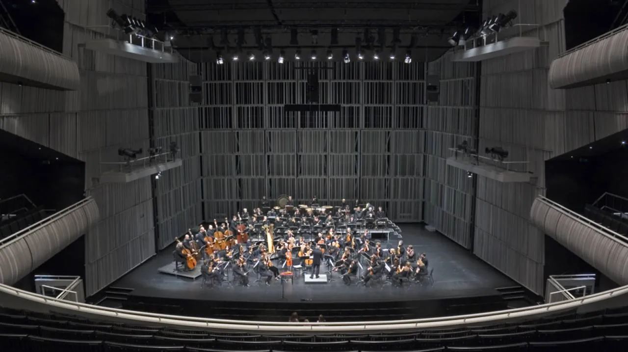 Vista interior amplia de una sala de conciertos con una orquesta completa actuando en el escenario, enmarcada por altos paneles acústicos grises y sistemas de iluminación superiores, con asientos escalonados visibles en primer plano.