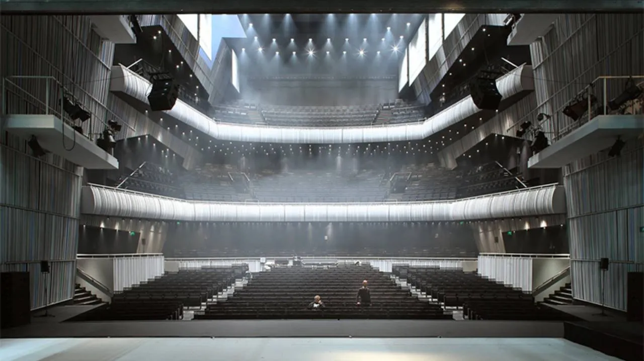 Vista interior de un gran auditorio de conciertos con asientos oscuros en niveles, múltiples balcones blancos curvados y una espectacular iluminación superior, visto desde el escenario hacia afuera.