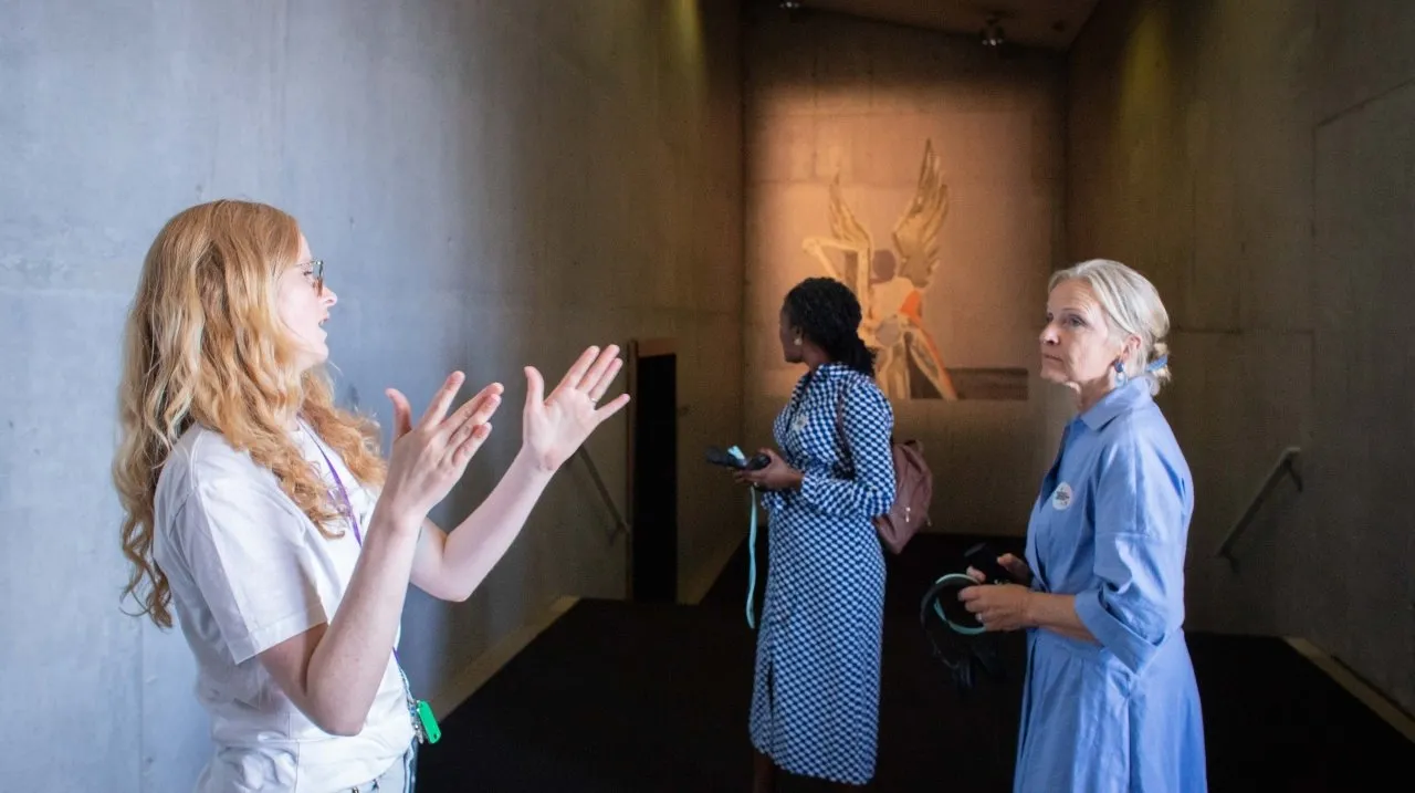Three women in a modern gallery space with concrete walls, one gesturing as a tour guide while two visitors listen, with artwork visible in the background