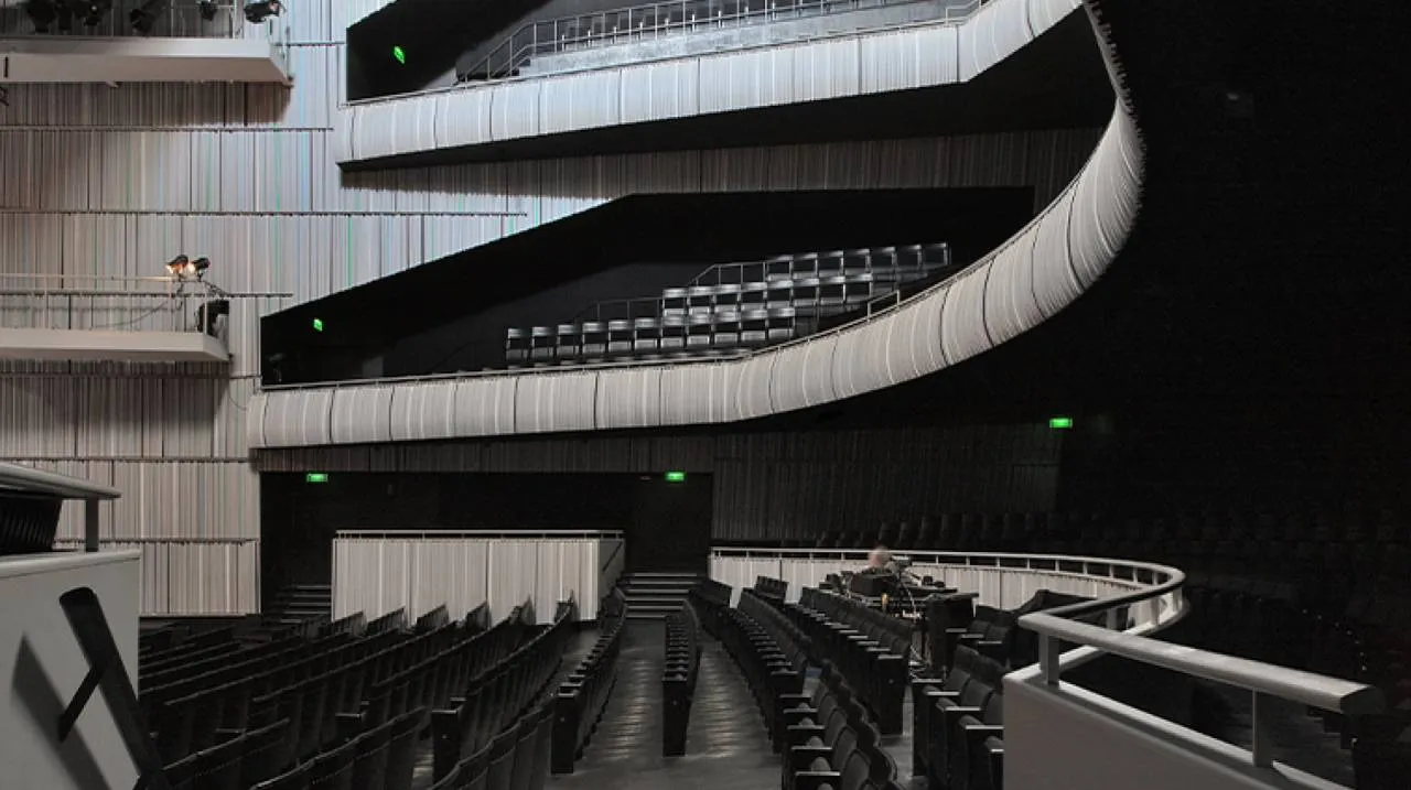 Vista interior de una moderna sala de conciertos con balcones blancos curvos y amplios, filas de asientos oscuros y luces de salida verdes visibles sobre un fondo oscuro.