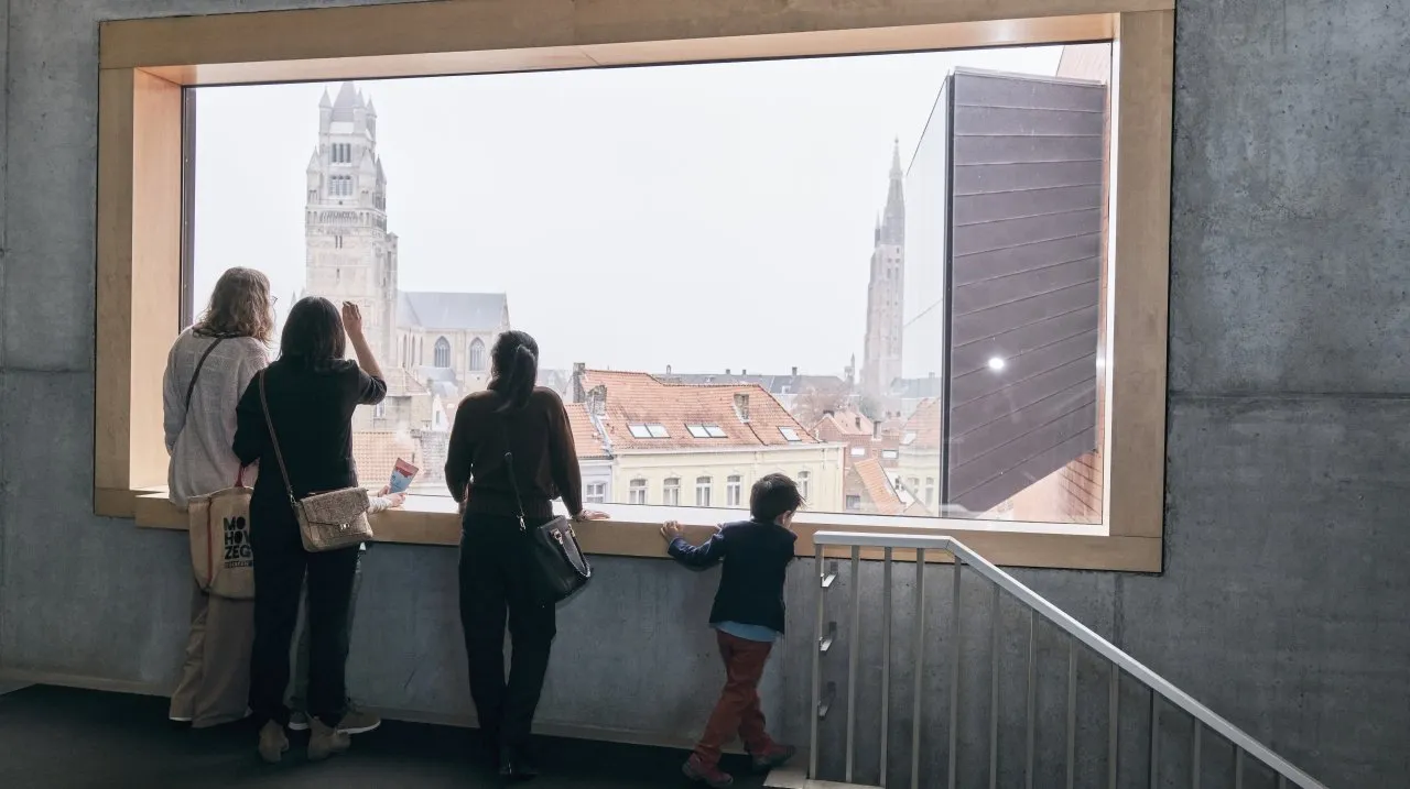 People standing inside looking through a large rectangular window at the Bruges cityscape with the Belfry tower and red rooftops visible outside