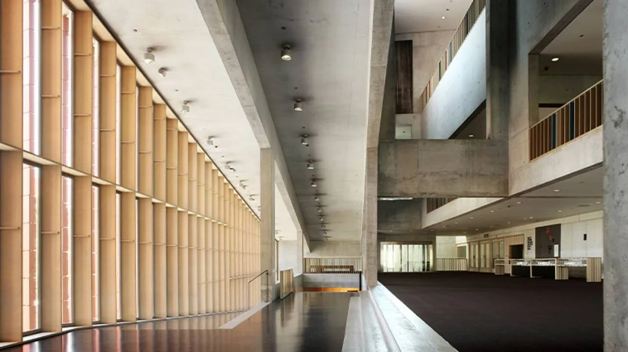 Largo pasillo interior con altas ventanas verticales a la izquierda y un atrio de hormigón de varios niveles a la derecha.