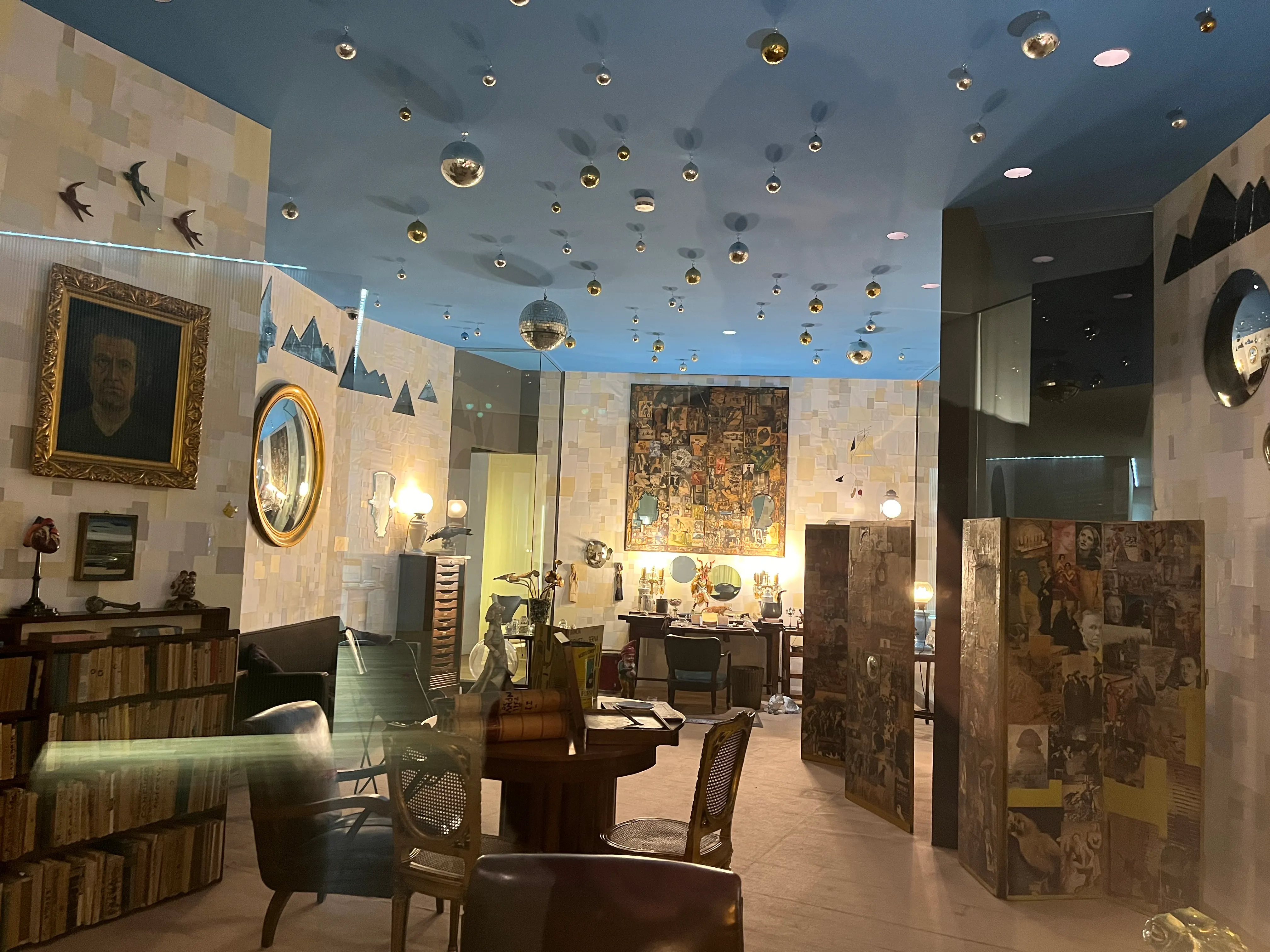 Interior de un museo con techo azul decorado con esferas metálicas, obras de arte enmarcadas y muebles