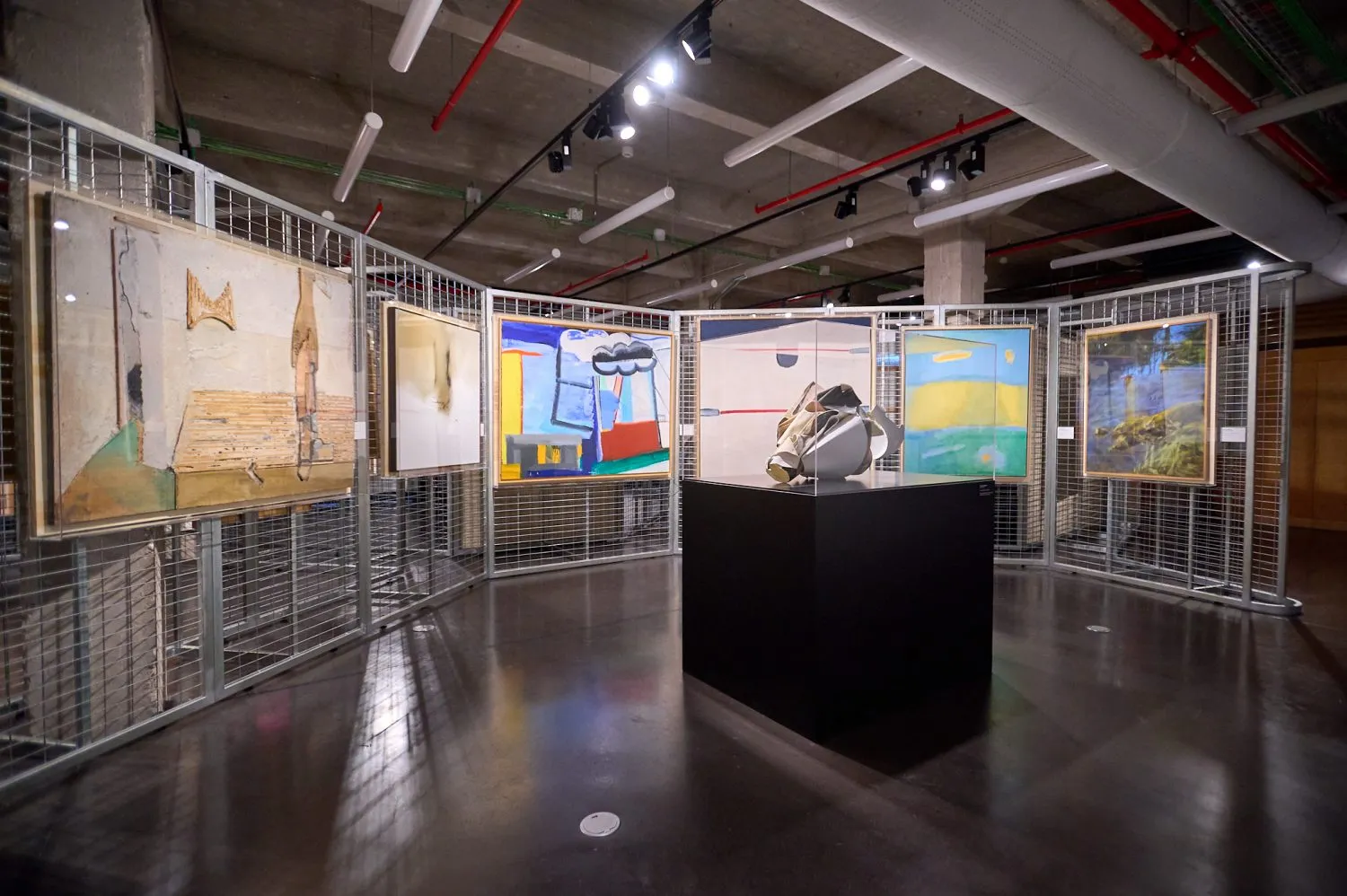 Obras de arte expuestas en una galería con vallas metálicas y un pedestal negro central bajo luces de techo de estilo industrial