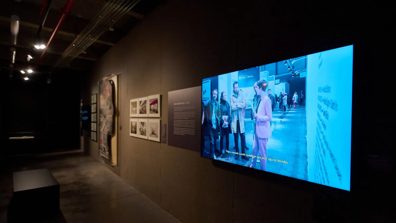 Duży ekran wyświetlający film z ludźmi w przestrzeni publicznej, zamontowany na ciemnej ścianie galerii z oprawionymi dziełami sztuki i panelami z tekstem w pobliżu