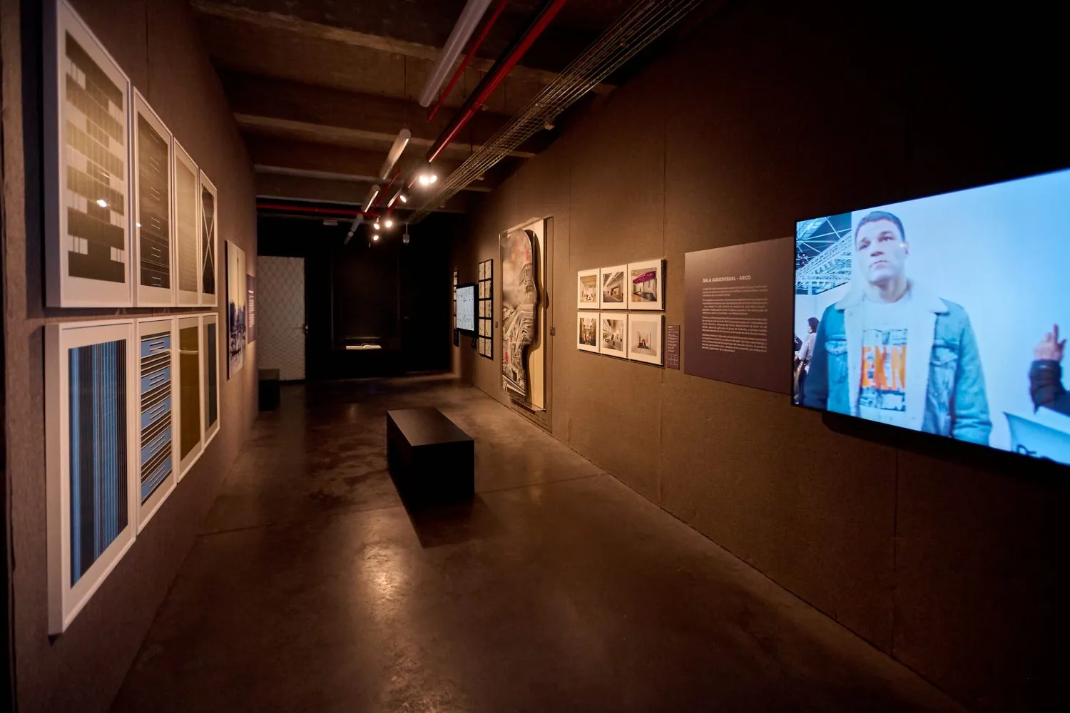 Pasillo de galería con obras de arte enmarcadas a la izquierda y una pantalla de video a la derecha