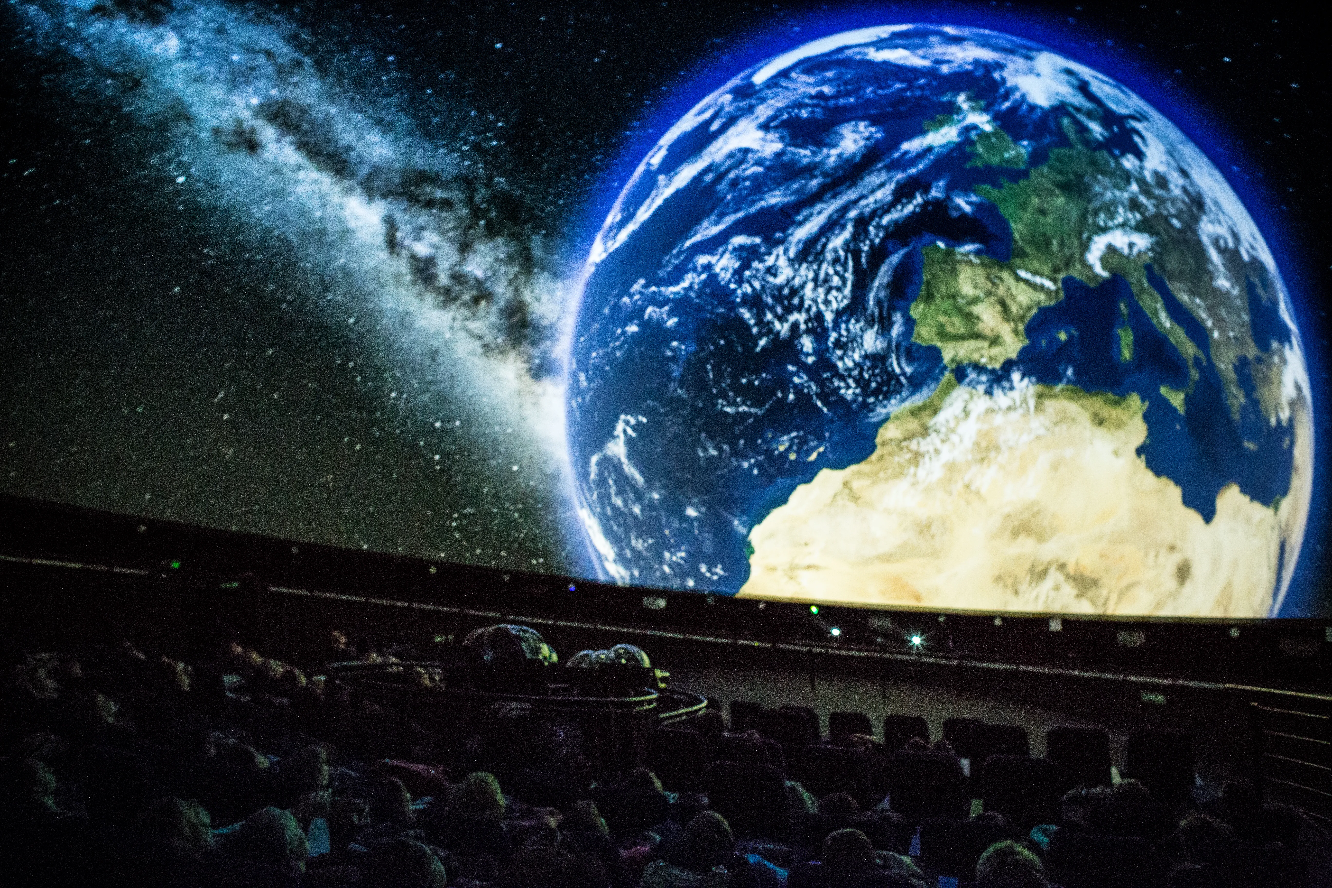 Widownia siedząca w ciemnym teatrze planetarium oglądająca dużą projekcję Ziemi i galaktyki Drogi Mlecznej na kopule.
