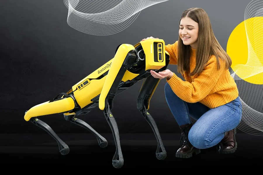 Kobieta w żółtym swetrze kuca obok żółtego robota-psa z czarnymi nogami i cyfrowym wyświetlaczem na grzbiecie.