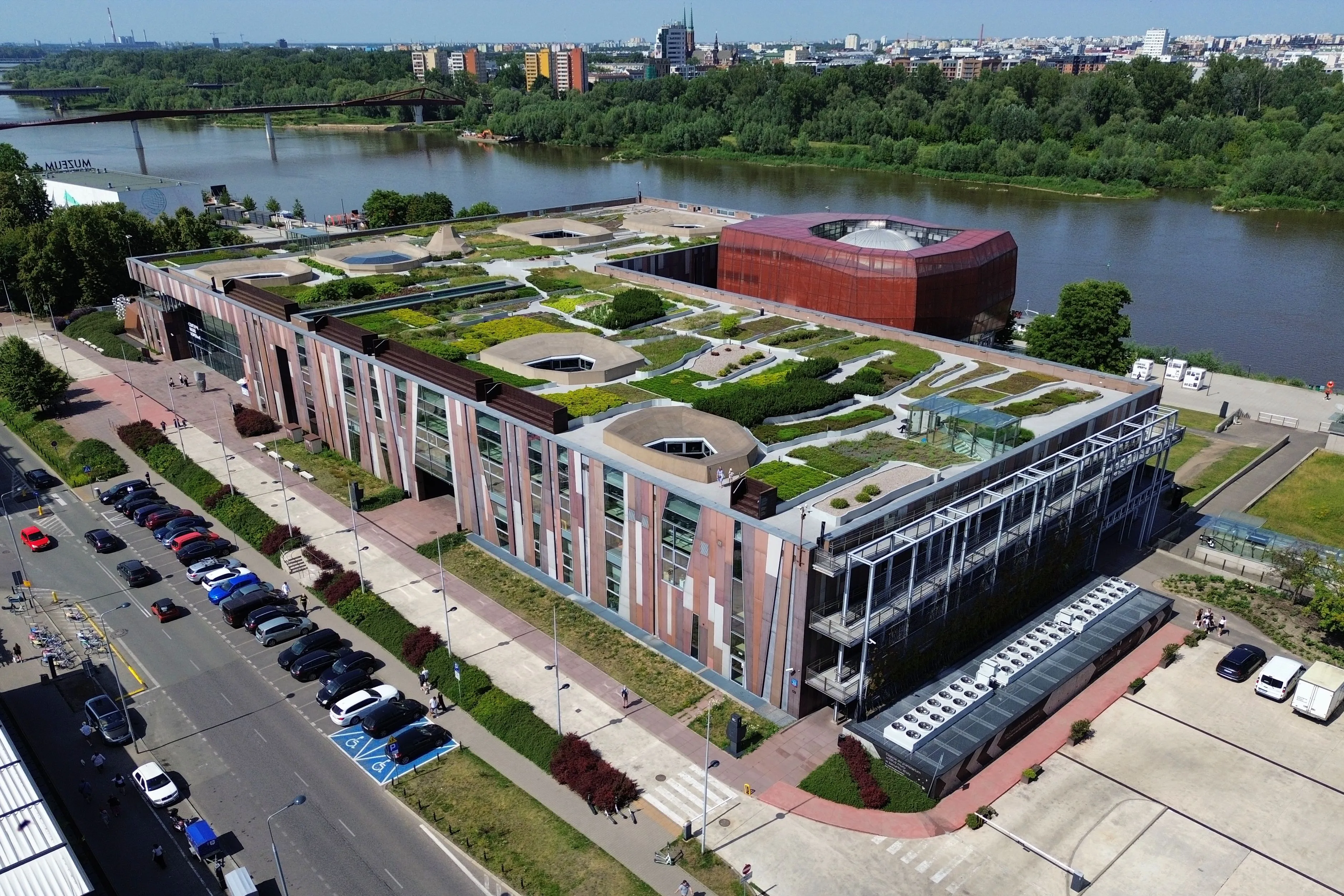 Widok z lotu ptaka na Centrum Nauki Kopernik z zielonym dachem, przylegające do rzeki i panoramy miasta.