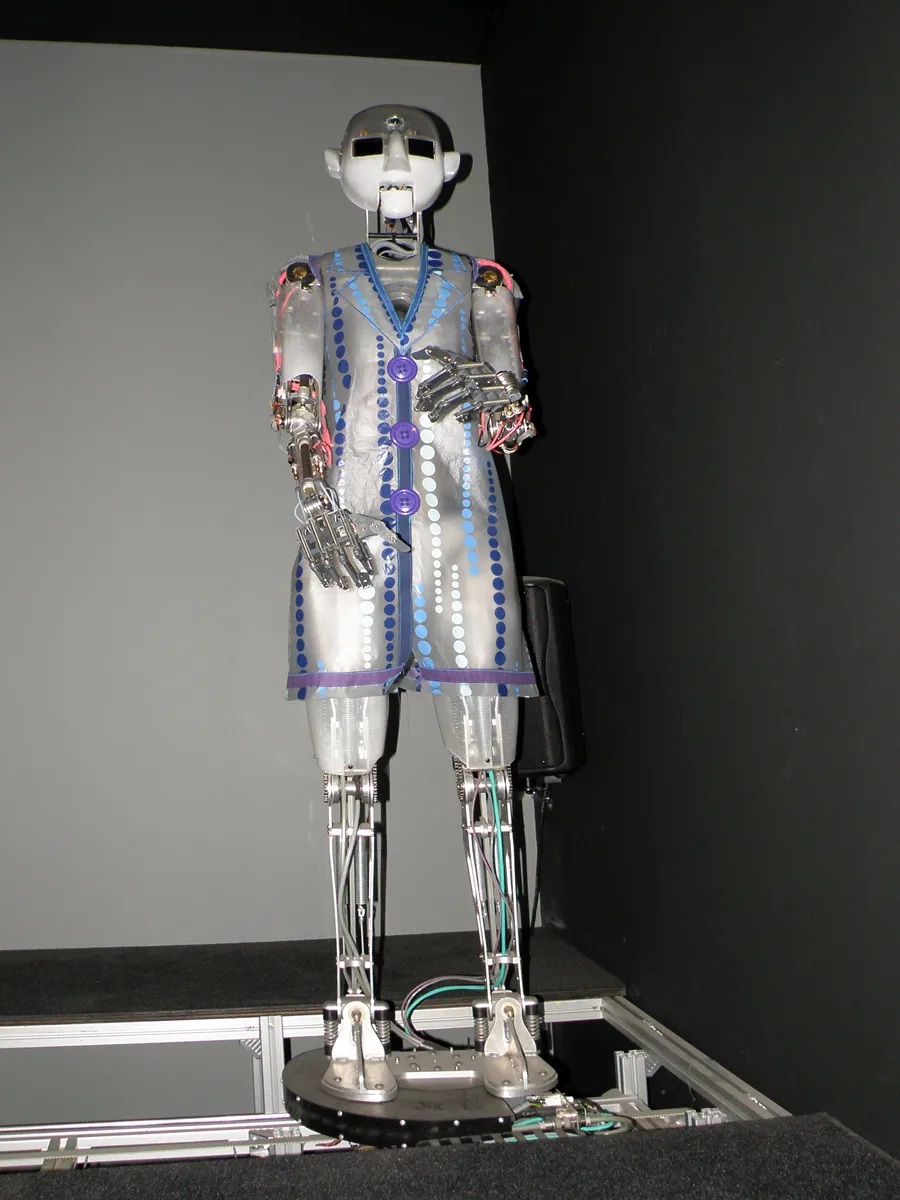 Un robot humanoide con un cuerpo transparente y partes mecánicas internas, de pie sobre una plataforma en una habitación oscura.