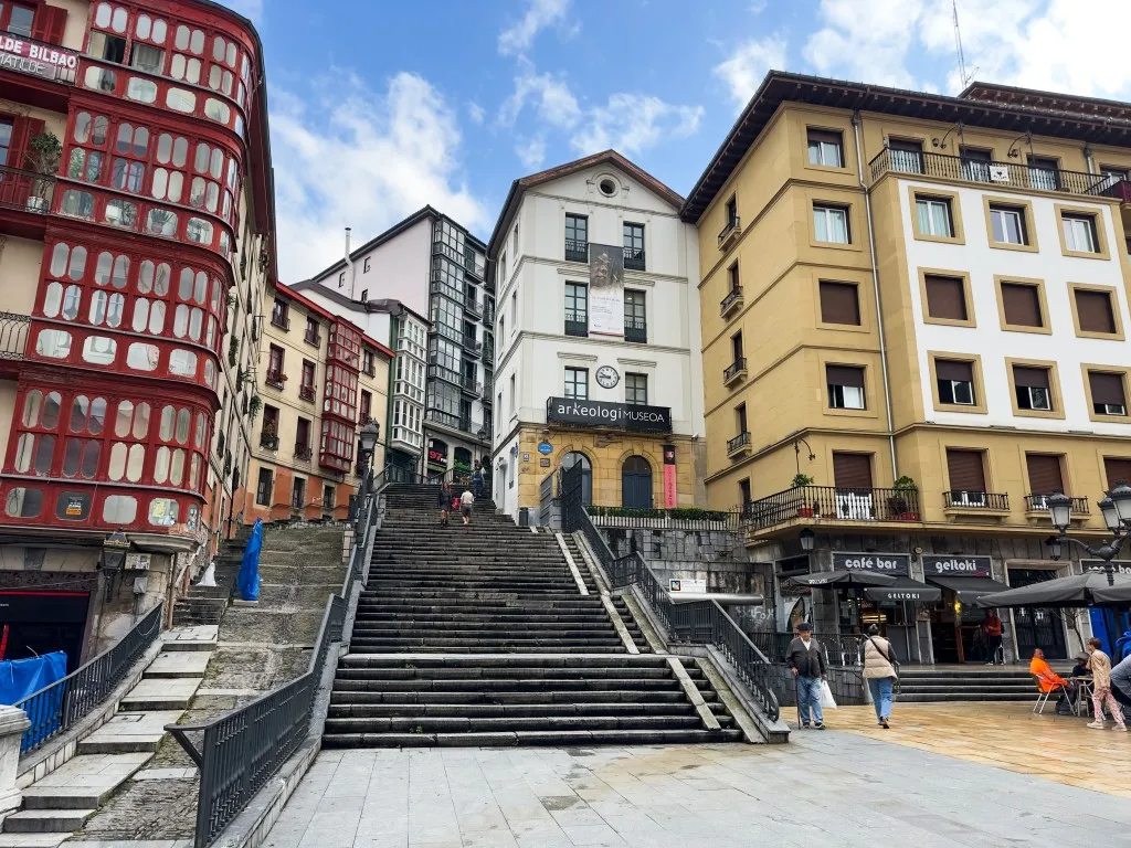 Amplia escalinata de piedra flanqueada por edificios coloridos en Bilbao, con un distintivo edificio rojo a la izquierda, un edificio central blanco y un edificio amarillo a la derecha, bajo un cielo parcialmente nublado con peatones en la base.