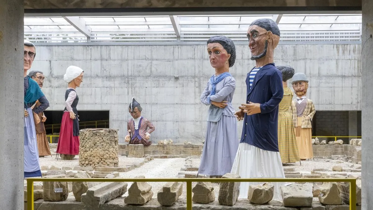 Grandes figuras de personas con vestimenta histórica situadas entre ruinas arqueológicas dentro de un sitio de excavación cubierto con techo de cristal y barandilla de seguridad amarilla.