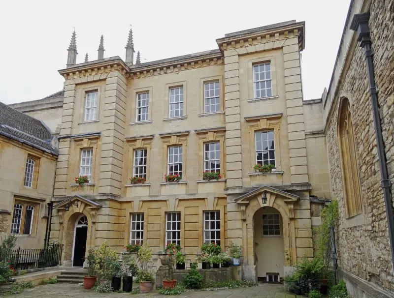 Stone facade of a historic college building with multiple windows, arched doorways, potted plants, and decorative pinnacles on the roofline