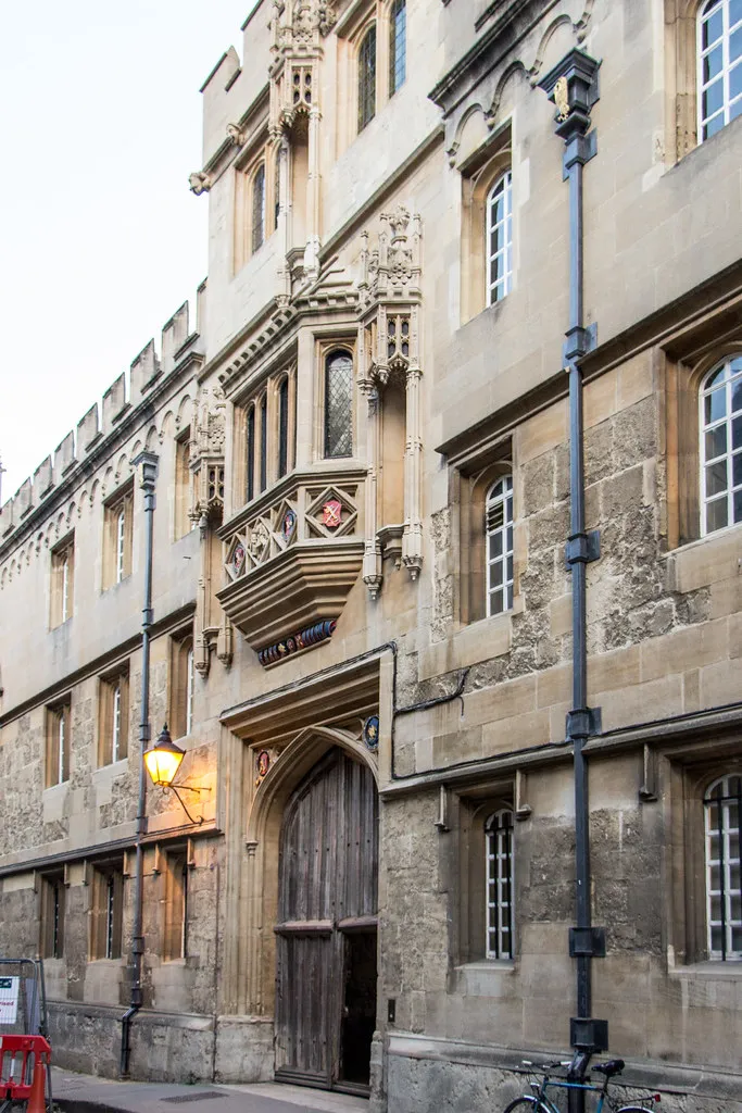 Stone facade of Corpus Christi College featuring a Gothic oriel window with coat of arms above a large arched wooden entrance door