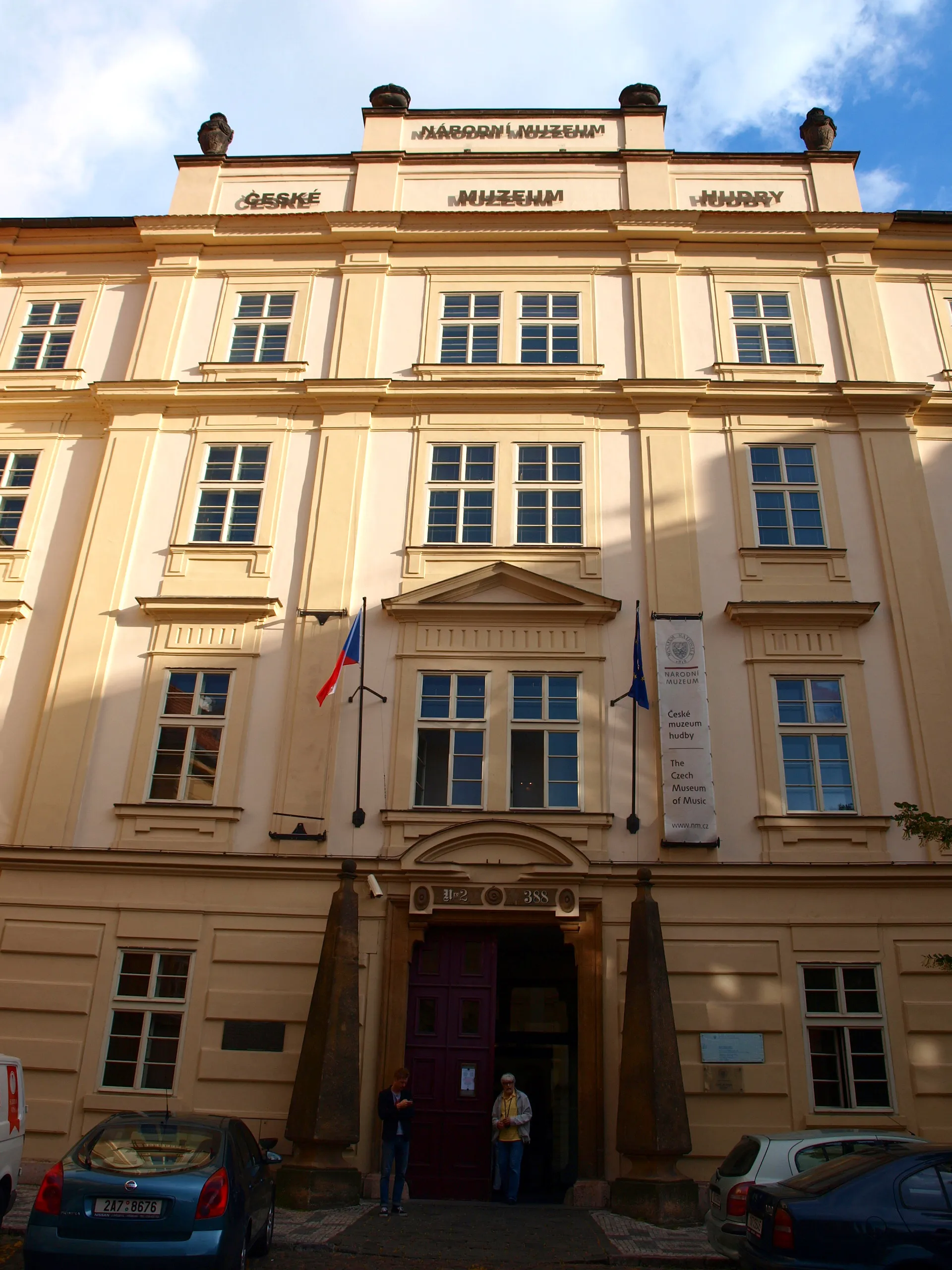 Beige barokke gebouw met meerdere ramen en een centrale ingang, met de tekst 'ČESKÉ MUZEUM HUDBY' boven de ingang en op de gevel, met twee vlaggen en mensen die bij de ingang staan.