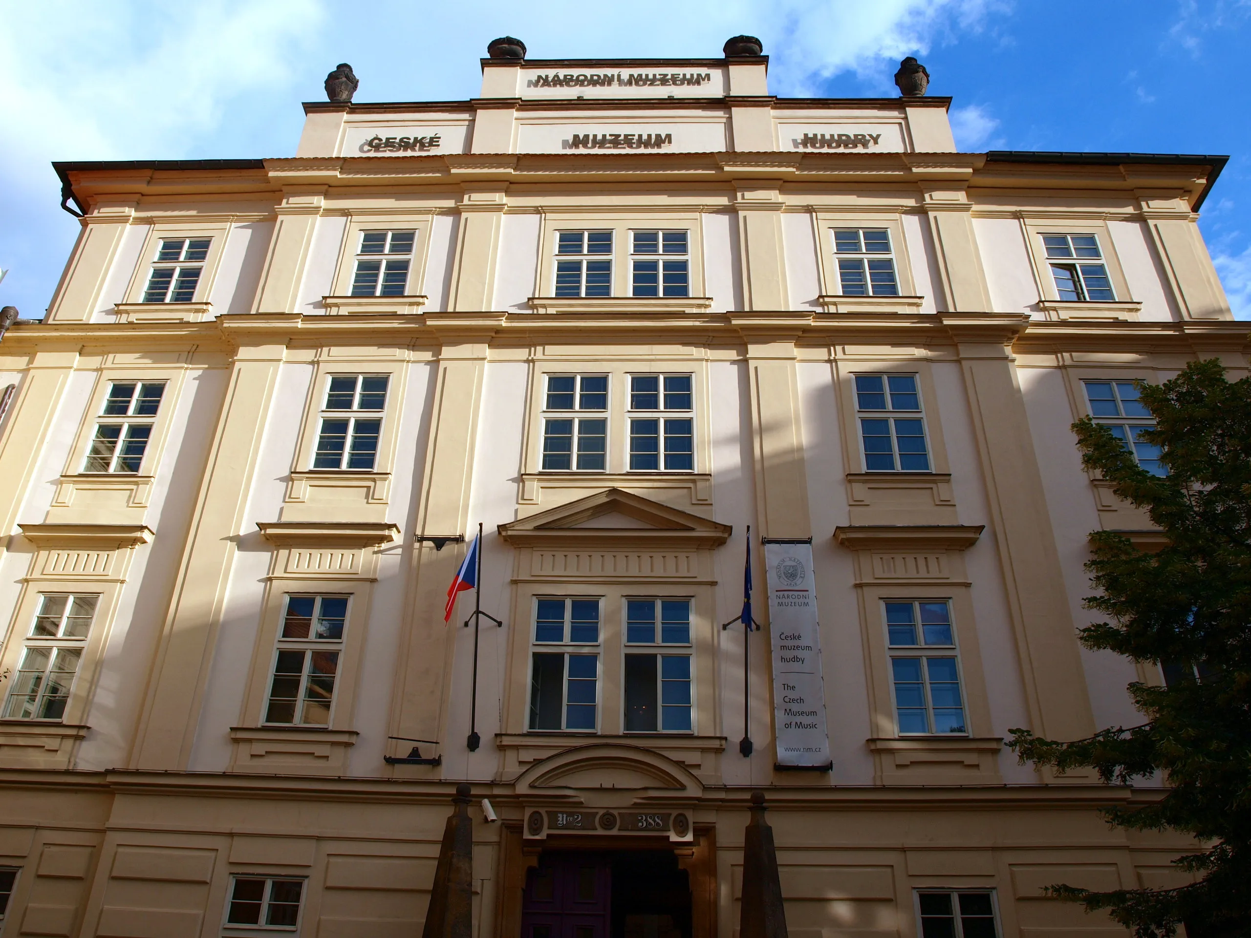 Beige barokke gebouw met meerdere ramen en een centrale ingang, met de naam 'ČESKÉ MUZEUM HUDBY' op de gevel onder een blauwe hemel.
