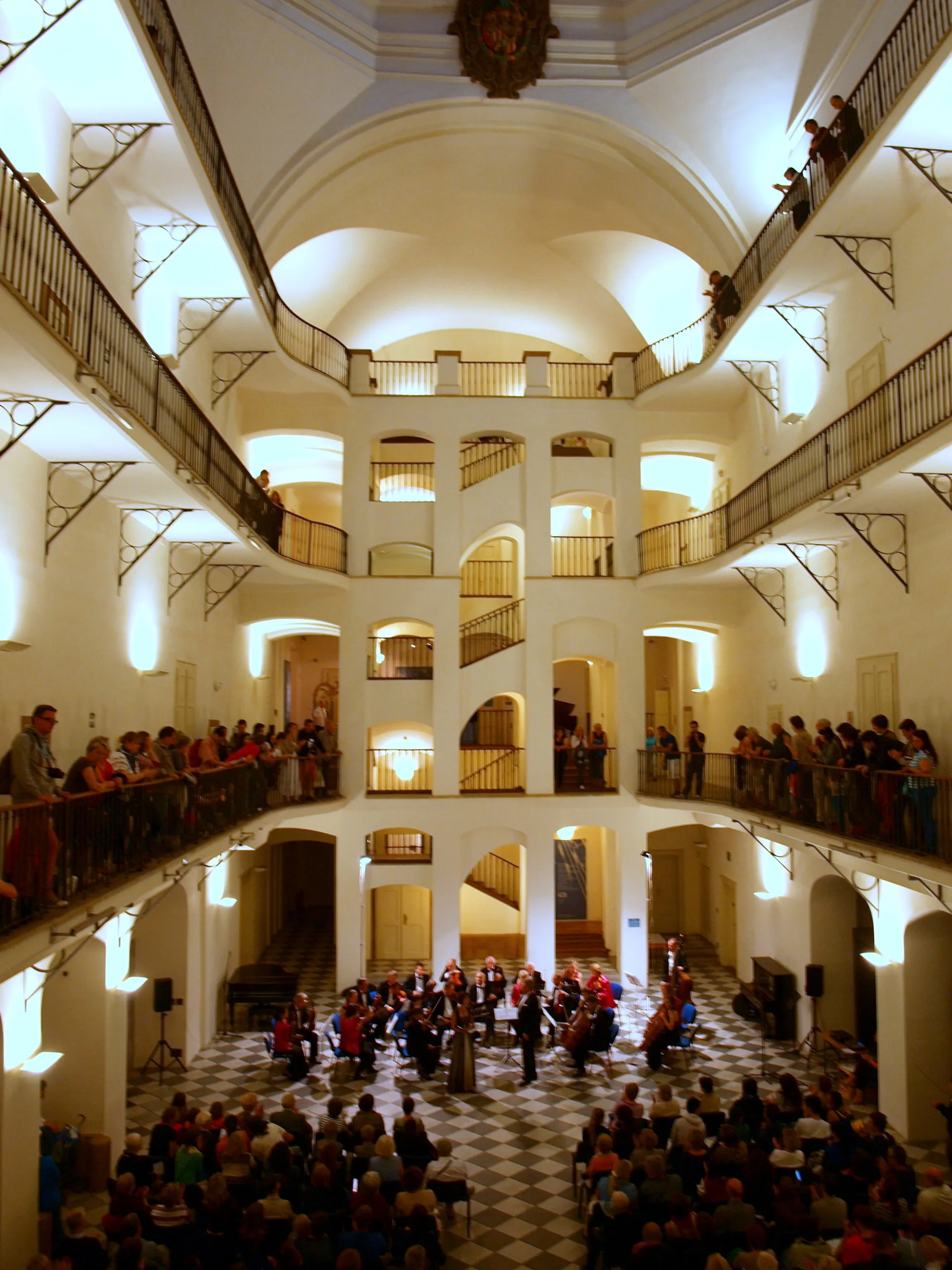 Interieur van een grote, meerlaagse atrium met witte muren, gebogen ramen en balkons, waar een muzikale uitvoering plaatsvindt met musici op het podium en publiek dat toekijkt.