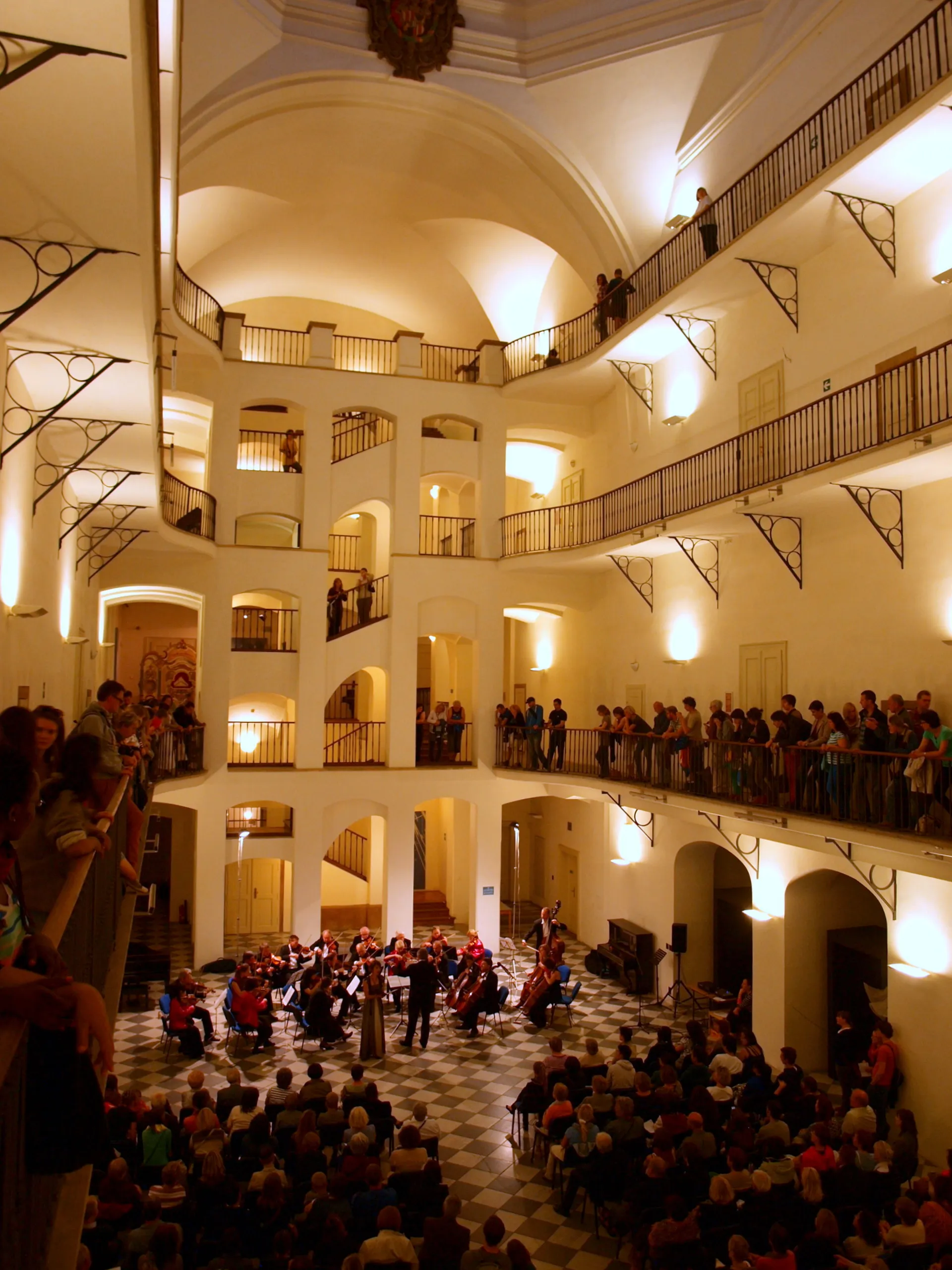 Interior of a grand hall with multiple levels, balconies, and a checkered floor, where a concert is in progress with an orchestra and audience.