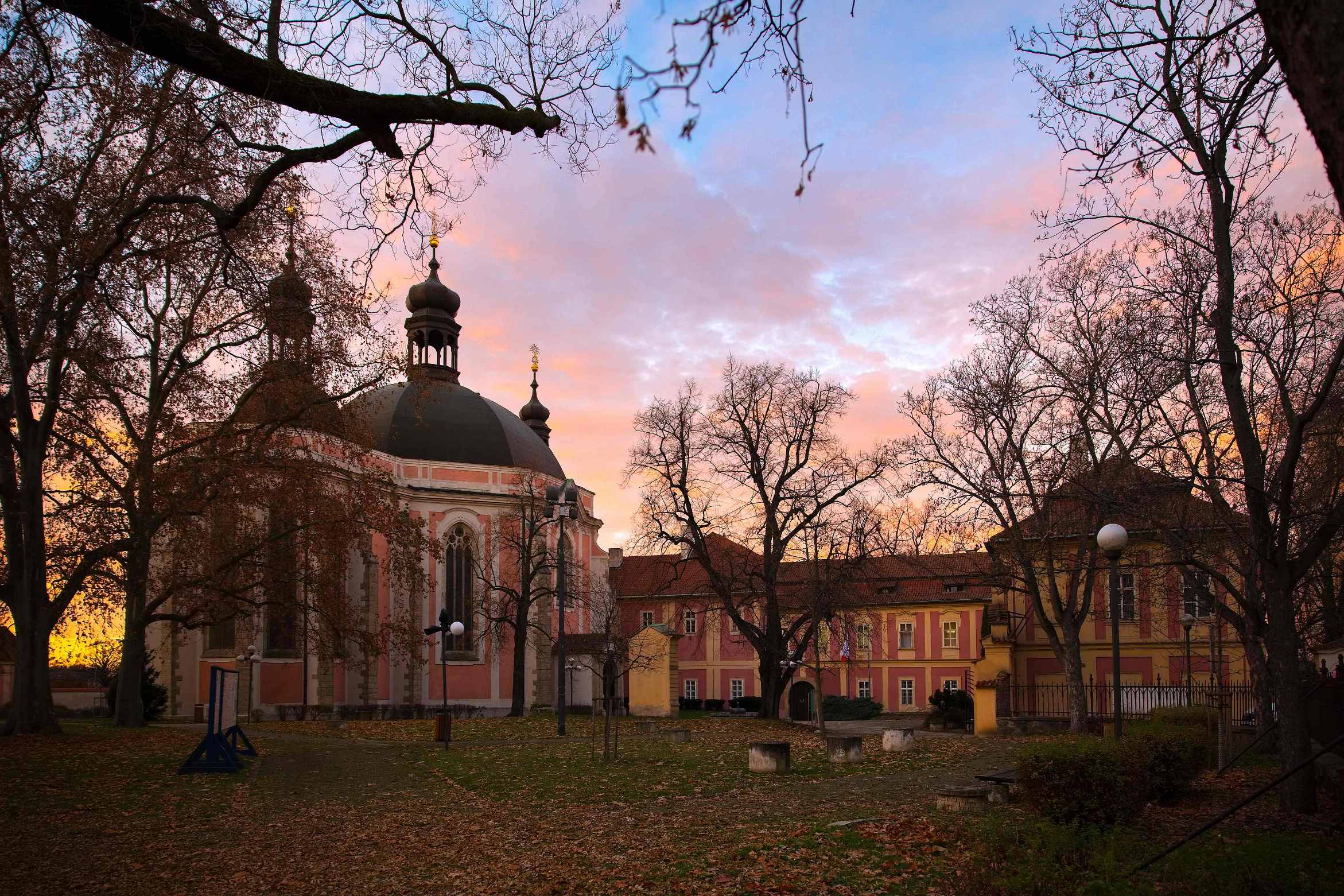 The Czech Police Museum building with a domed church and surrounding structures under a sunset sky with bare trees in the foreground