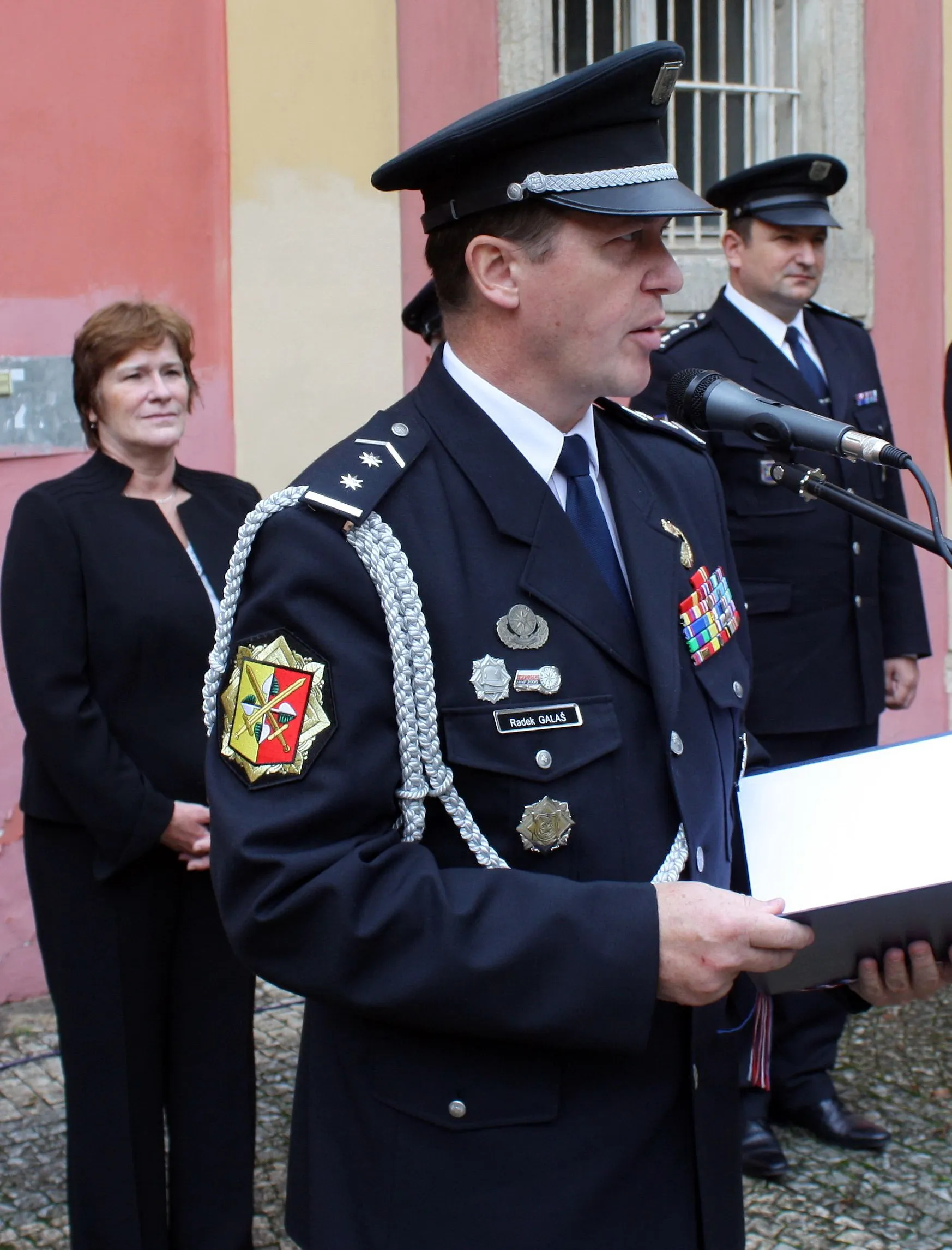 A police officer in a dark uniform with medals and insignia speaks at a microphone in front of a building with pink and beige walls.