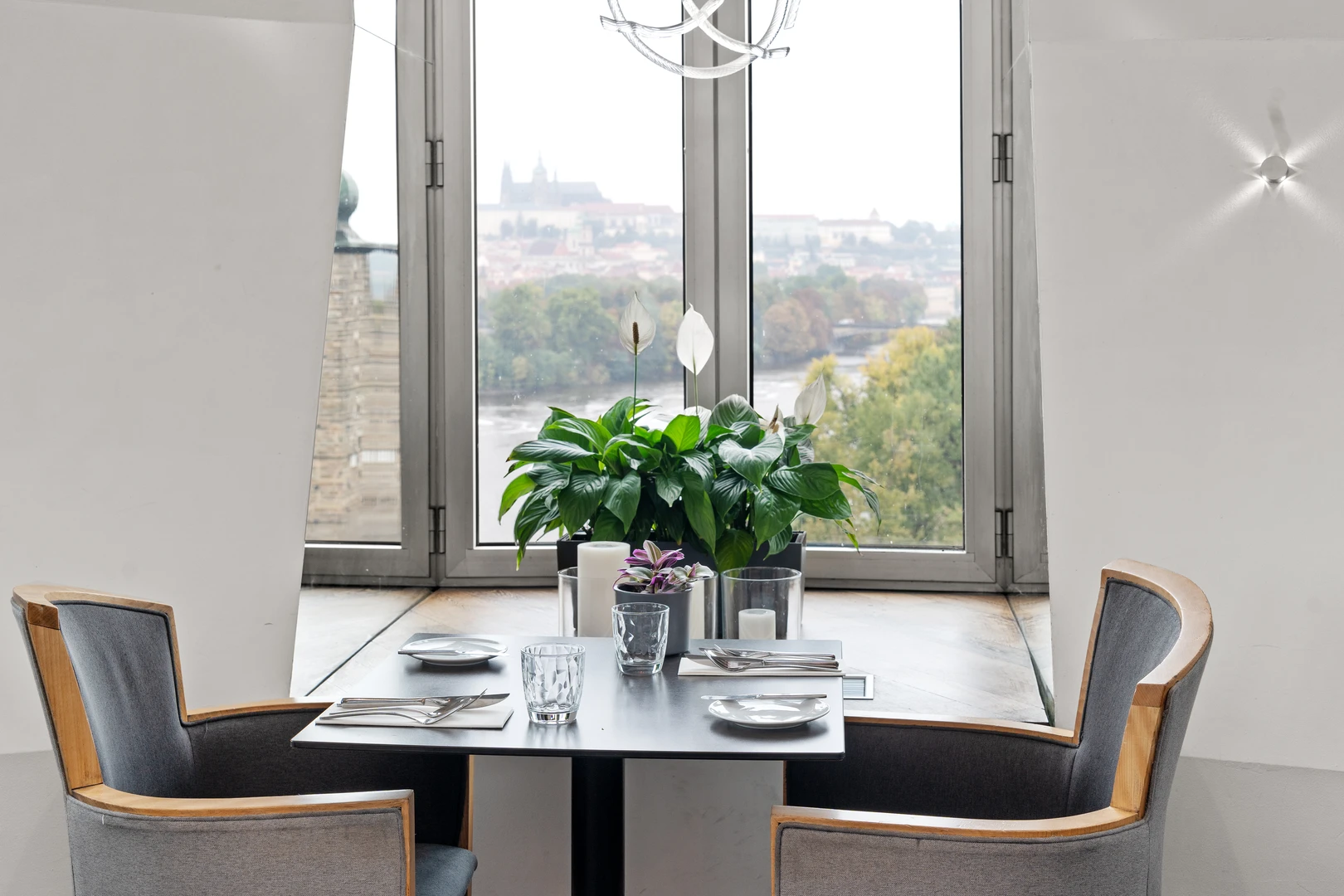 Una mesa de comedor con dos sillas y una planta en maceta, con vistas a un paisaje urbano a través de grandes ventanales.